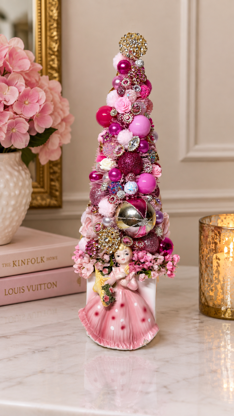 A decorative pink Christmas tree ornament featuring a small ballerina figurine at the base, embellished with pink and metallic ornaments, beads, and flowers, displayed on a marble surface next to a lit candle and pink flowers.