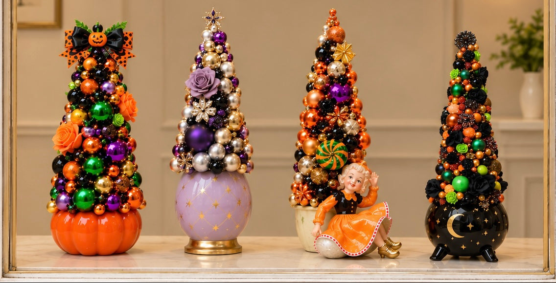 Four decorative Halloween-themed miniature Christmas trees in vibrant colors, displayed on a shelf under a gold 'Fleur Fancier' brand banner. The trees are adorned with colorful ornaments, beads, and faux foliage, with two decorated on pumpkins and one featuring a small angel figurine.