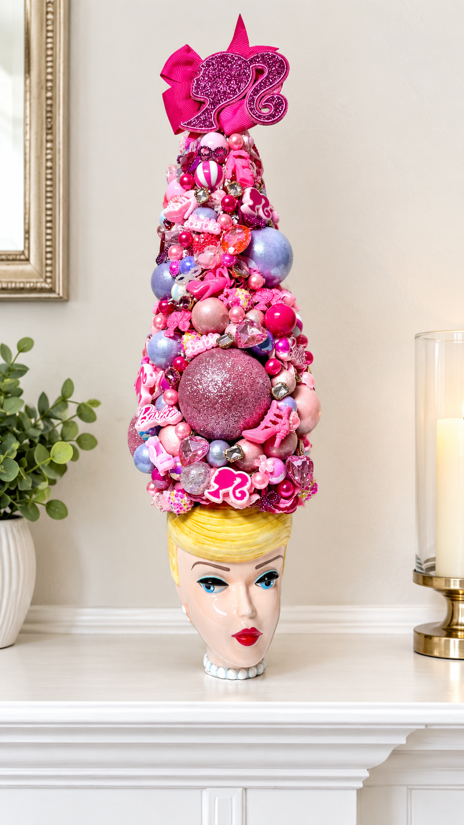 A decorative pink Barbie-themed Christmas tree crafted from ornaments, candies, and pink embellishments, displayed on a white mantel above a mannequin head with blonde hair and red lips.