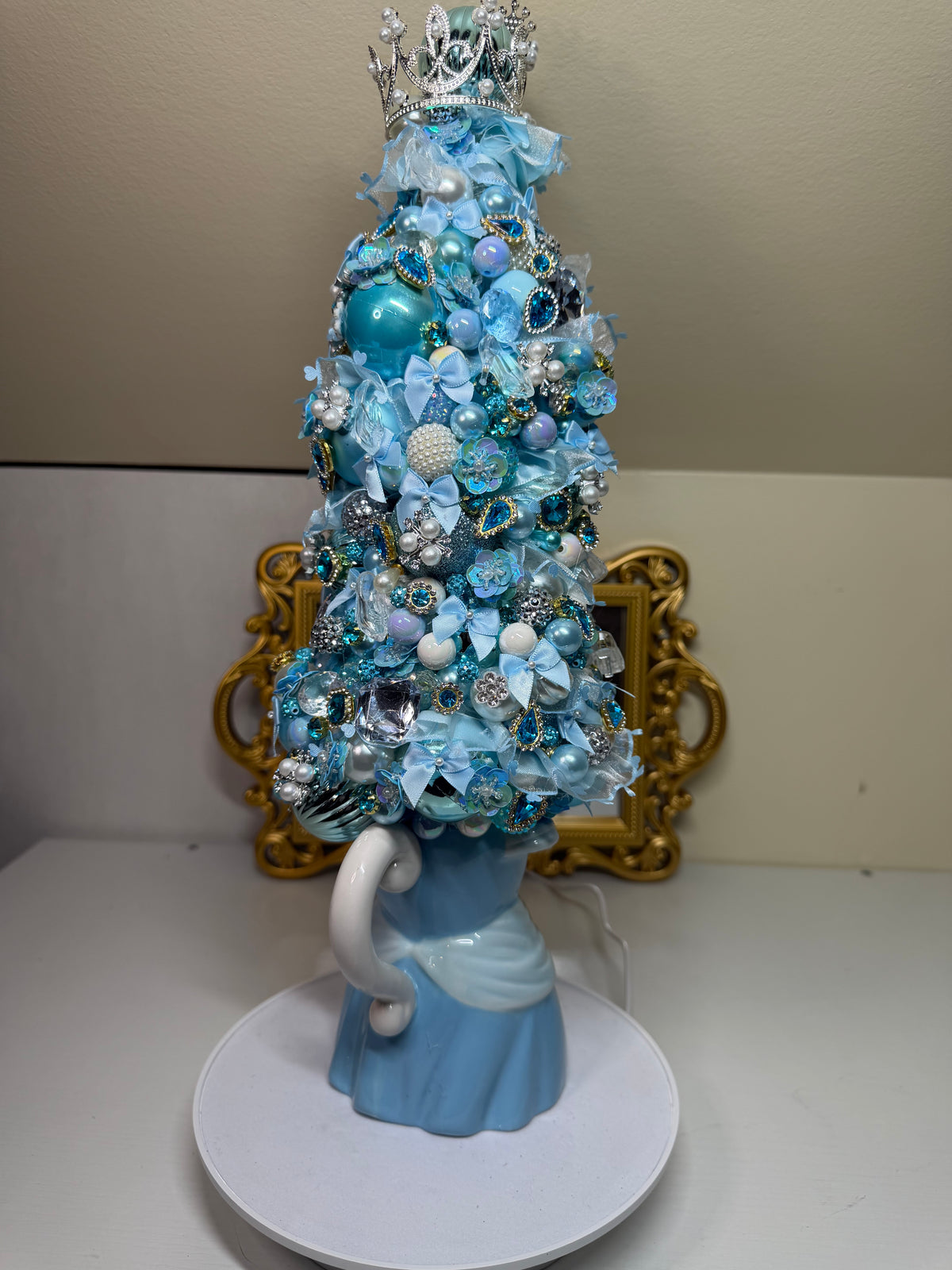 A decorative miniature Christmas tree in shades of light blue, turquoise, and silver, styled with ornaments, bows, and a silver crown, mounted on a light blue porcelain figure of a swan on a white circular base.