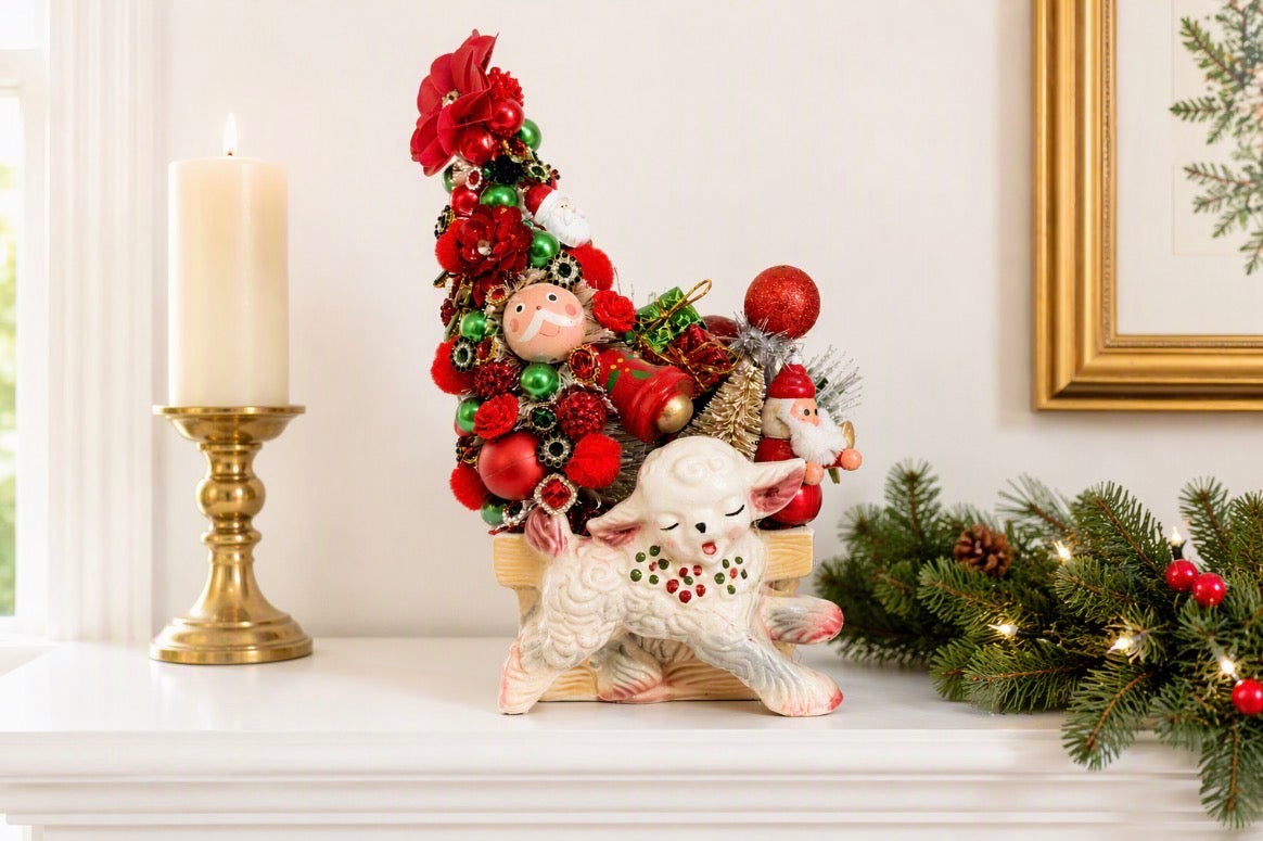 A festive Christmas ornament arrangement displayed on a white mantel, featuring a tall red Christmas tree with ornaments, a white lamb figurine, a gold candlestick, and holiday greenery.