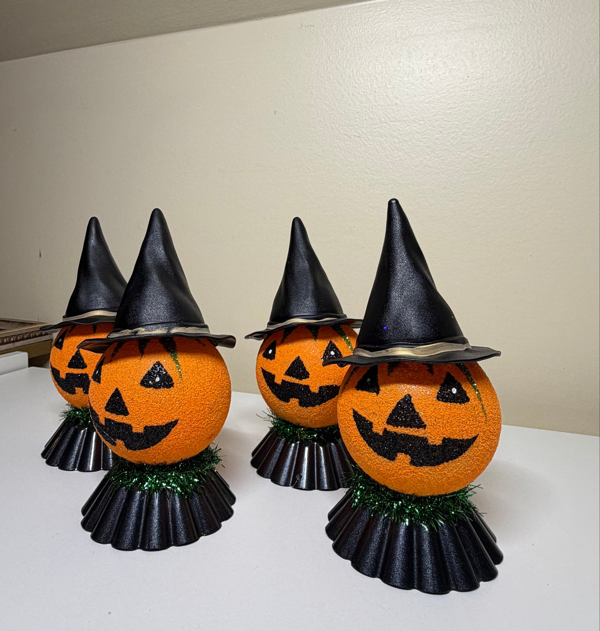 Four orange jack-o'-lantern pumpkins decorated with black witch hats and black pleated skirts, arranged on a white shelf against a beige wall.