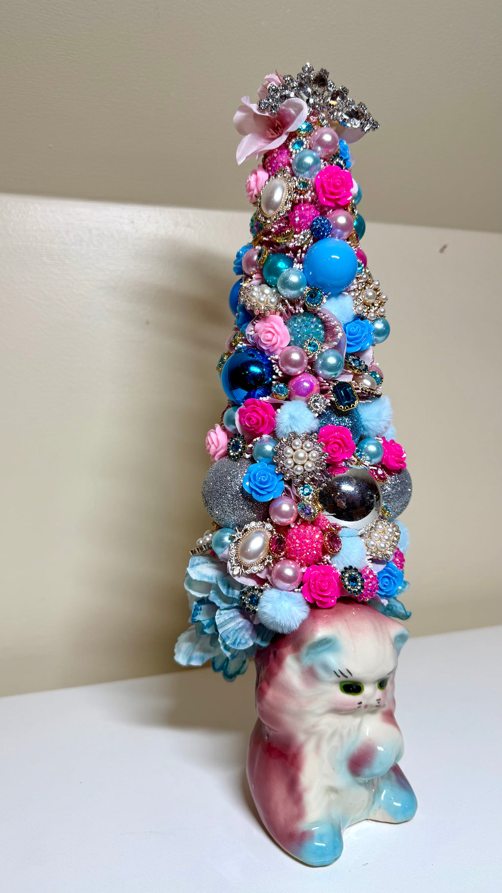 A whimsical, tall Christmas tree decoration made from colorful faux flowers, rhinestones, pearls, and blue ornaments, perched on top of a pink and blue cat figurine.