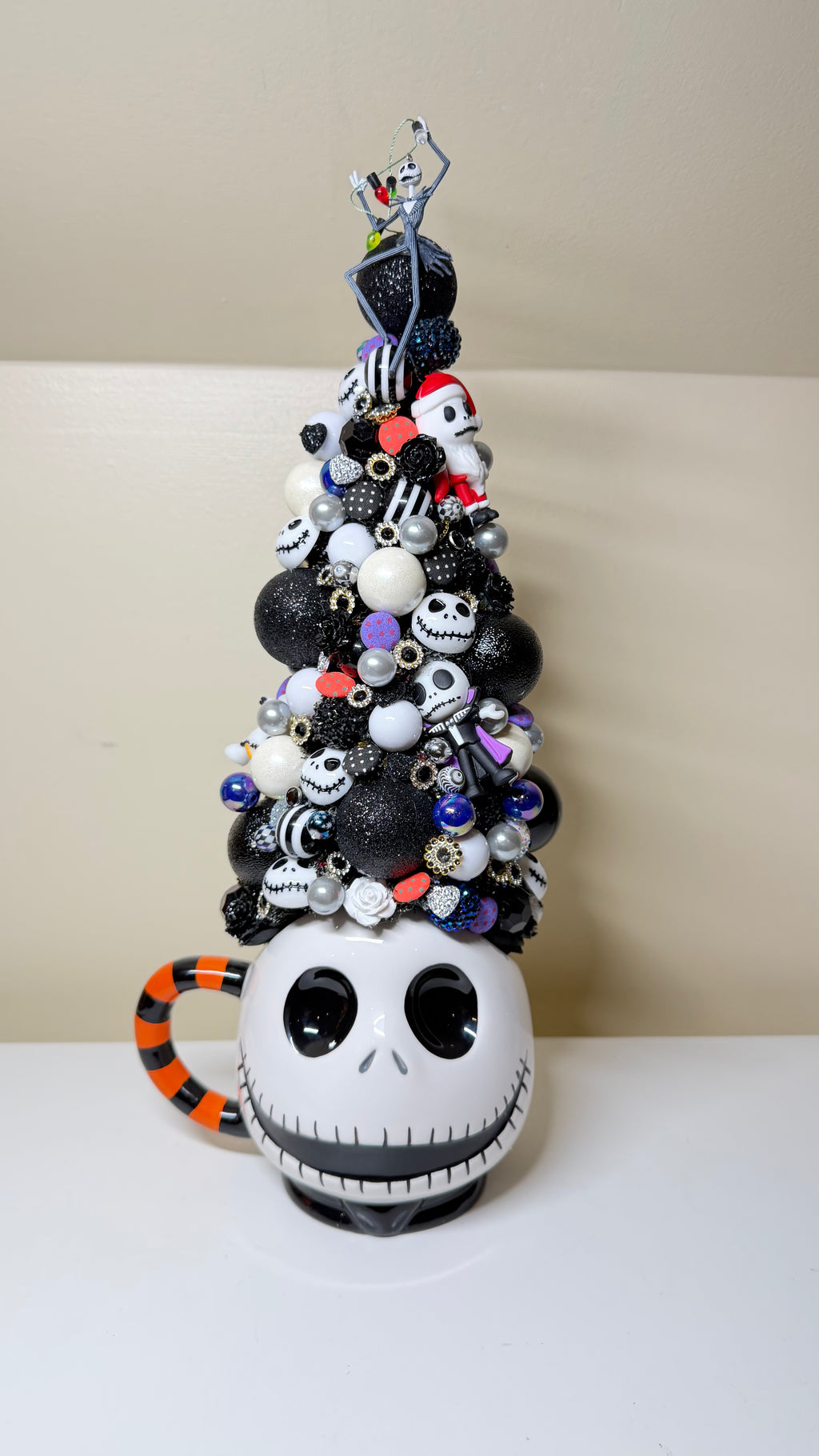 A Jack Skellington themed Christmas tree decoration shaped like a mug, featuring a white skull face, orange and black striped handle, and a tall tree of black, white, and silver ornaments topped with a small figurine.