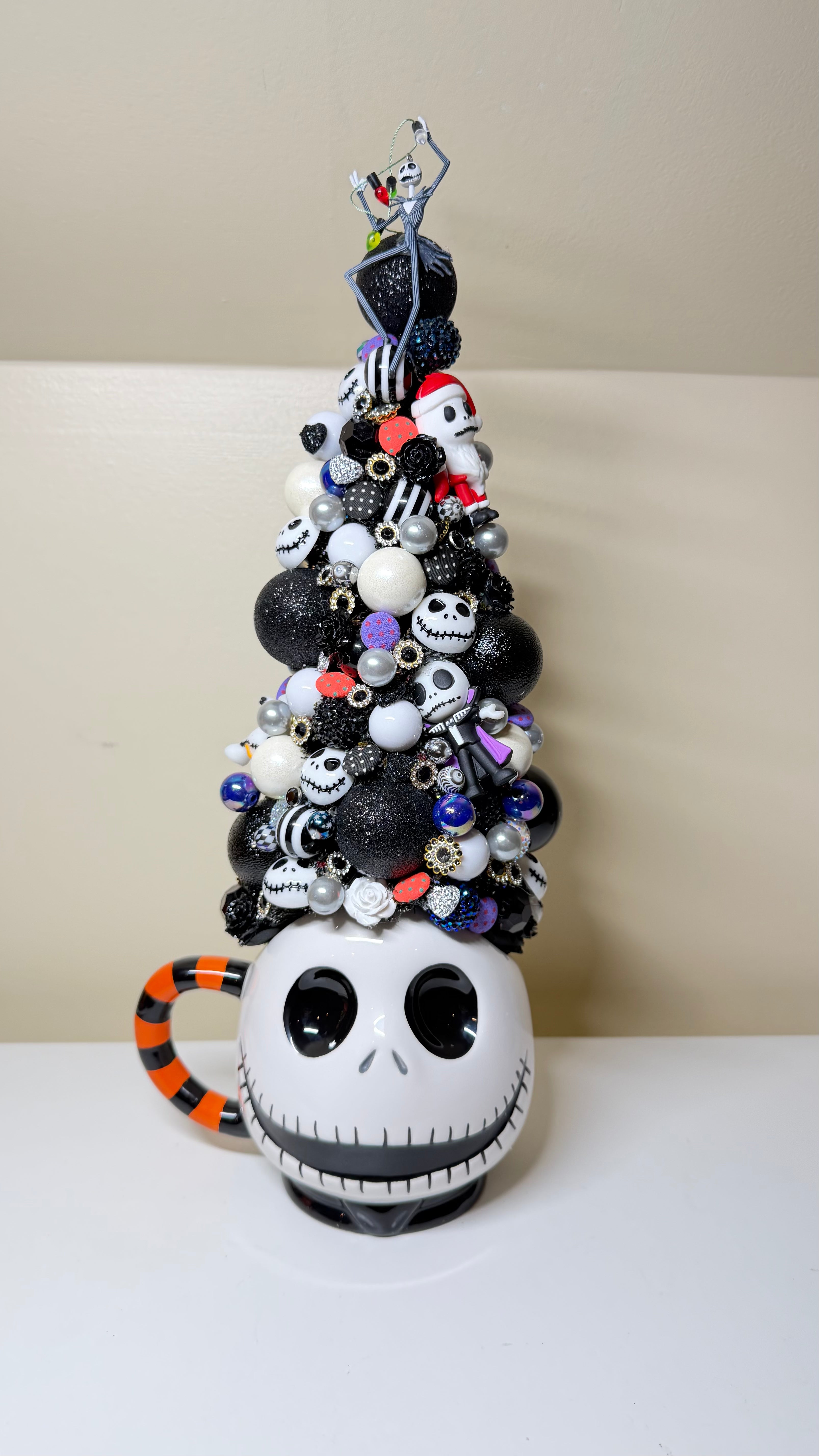 A Jack Skellington themed Christmas tree decoration shaped like a mug, featuring a white skull face, orange and black striped handle, and a tall tree of black, white, and silver ornaments topped with a small figurine.