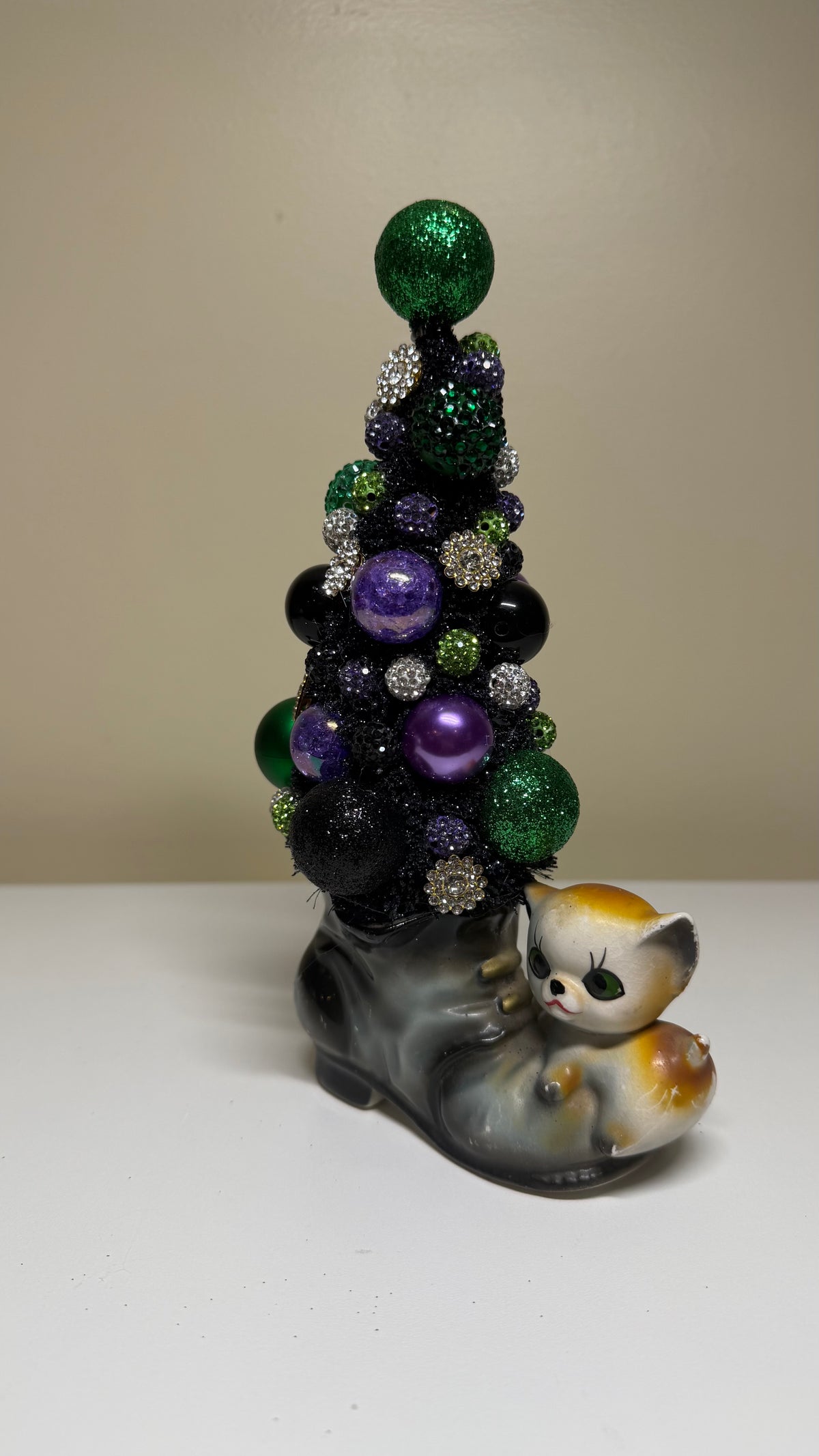A decorative Mardi Gras-themed ornament featuring a small black Christmas tree with green, purple, and silver ornaments, perched on a gray boot-shaped base with a white, orange, and black kitten figurine at the front, set against a plain beige wall and white surface.