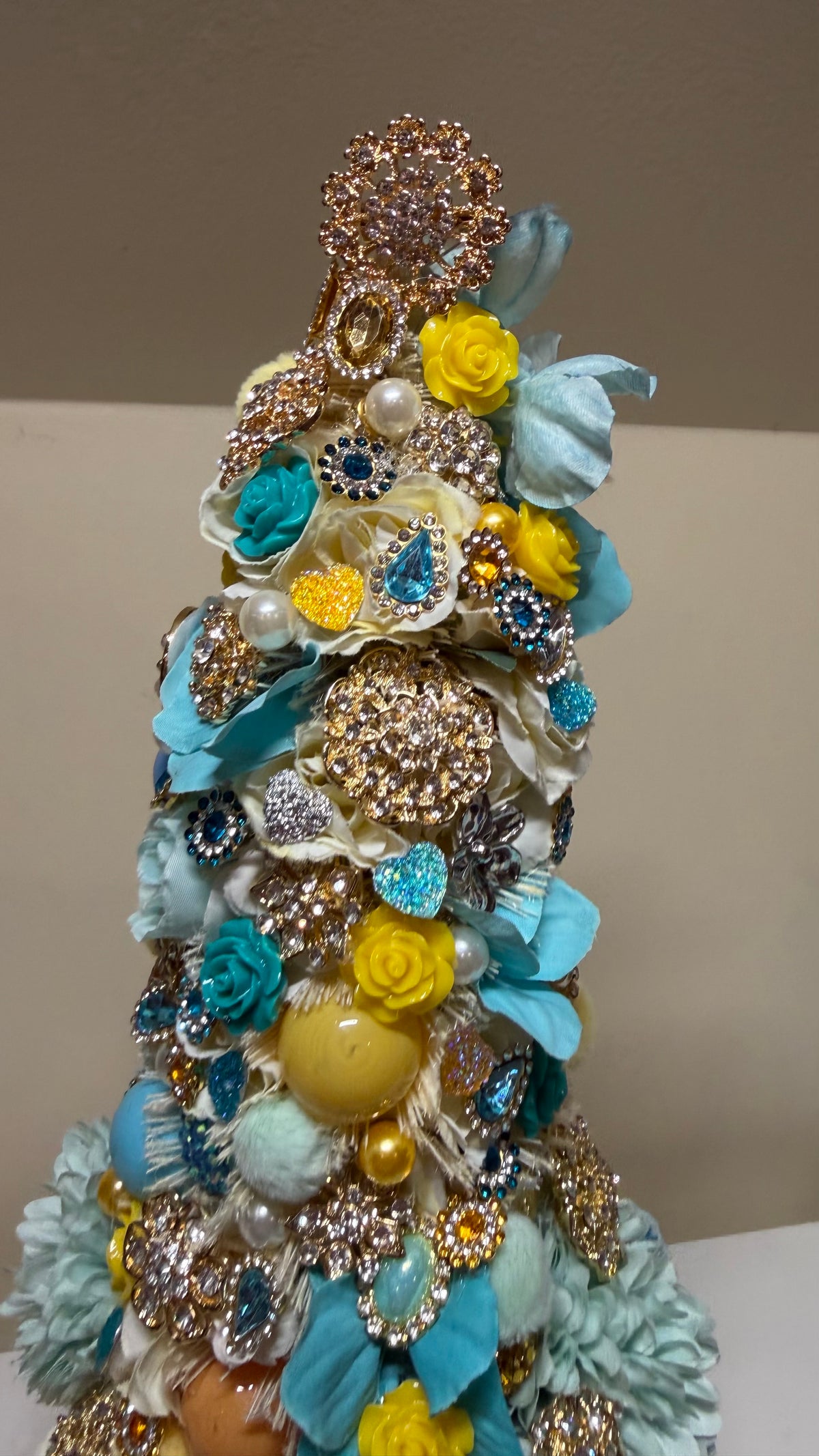 A decorative, tiered tree crafted from layered fashion jewelry, brooches, pearls, and faux flowers in light blue, yellow, gold, and silver, set against a plain beige background.