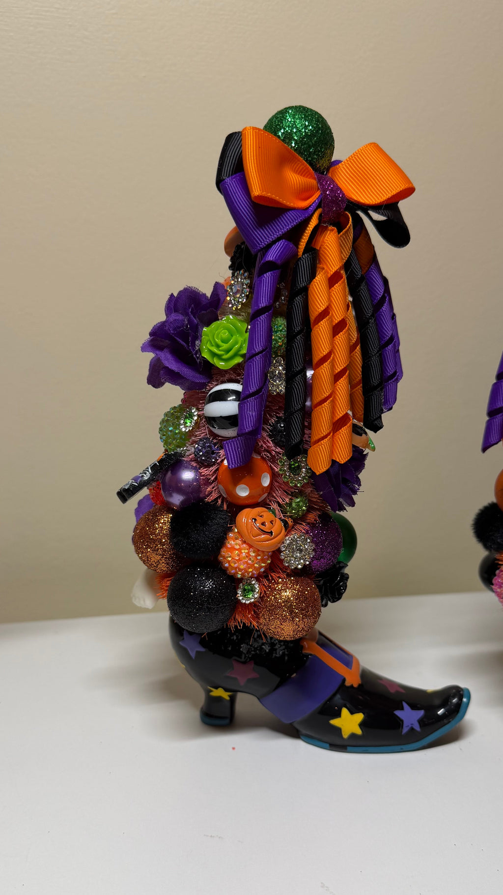 A Halloween-themed decorative boot figurine decorated with purple and orange ribbons, glittery pumpkins, colorful beads, and a purple bow, standing on a white surface against a beige wall.