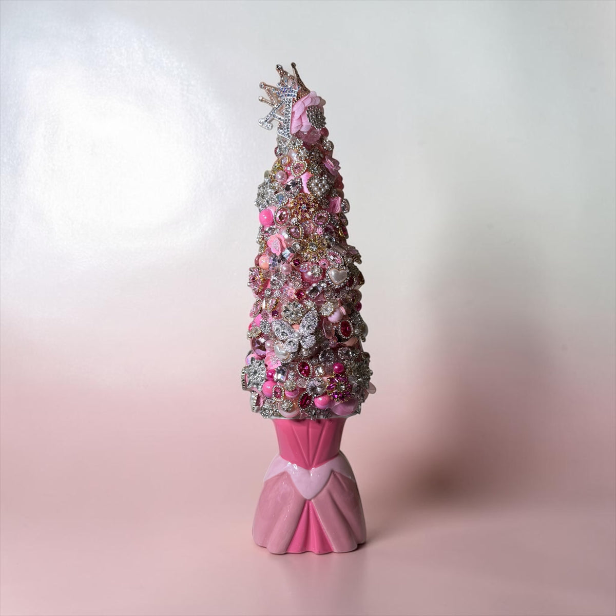 A decorative pink Christmas tree sculpture covered in pink and silver rhinestones, flowers, and embellishments, standing on a pink pedestal against a soft pink and white gradient background.