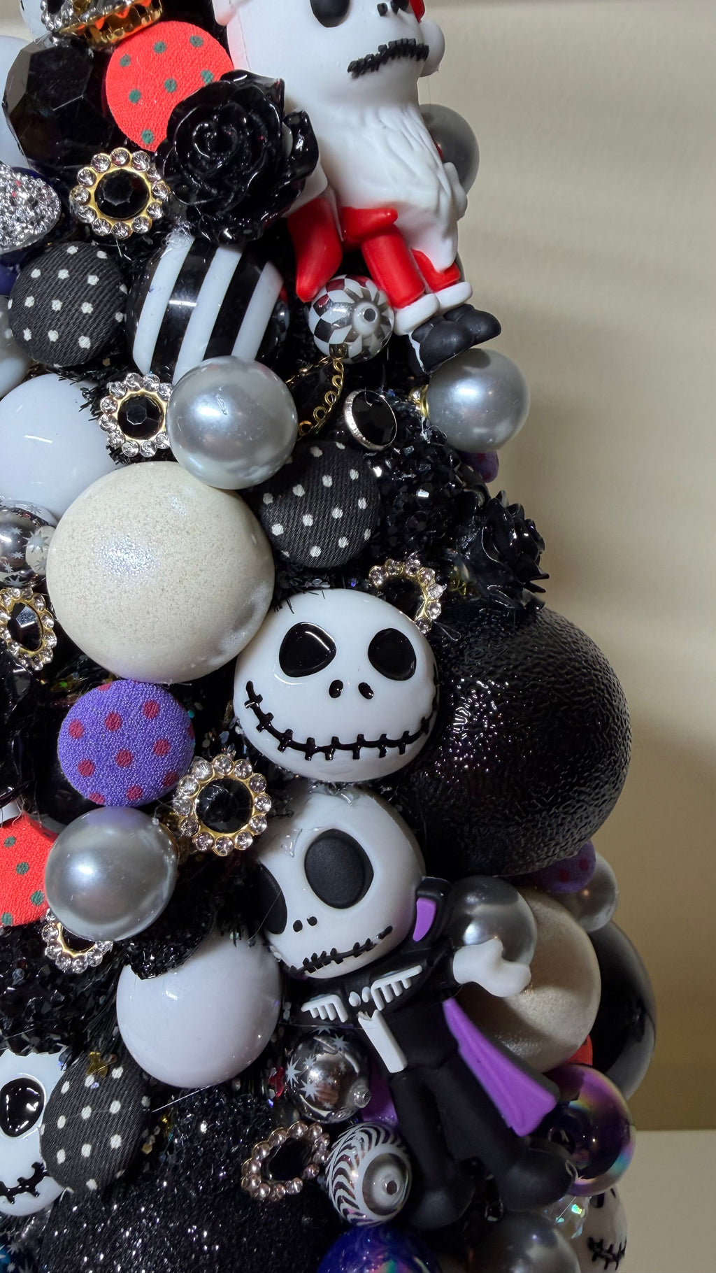 A close-up of a decorative Christmas tree covered in black, white, and silver ornaments, including skull-shaped baubles, striped pom-poms, pearl-like spheres, and small figurines, photographed indoors against a plain light background.