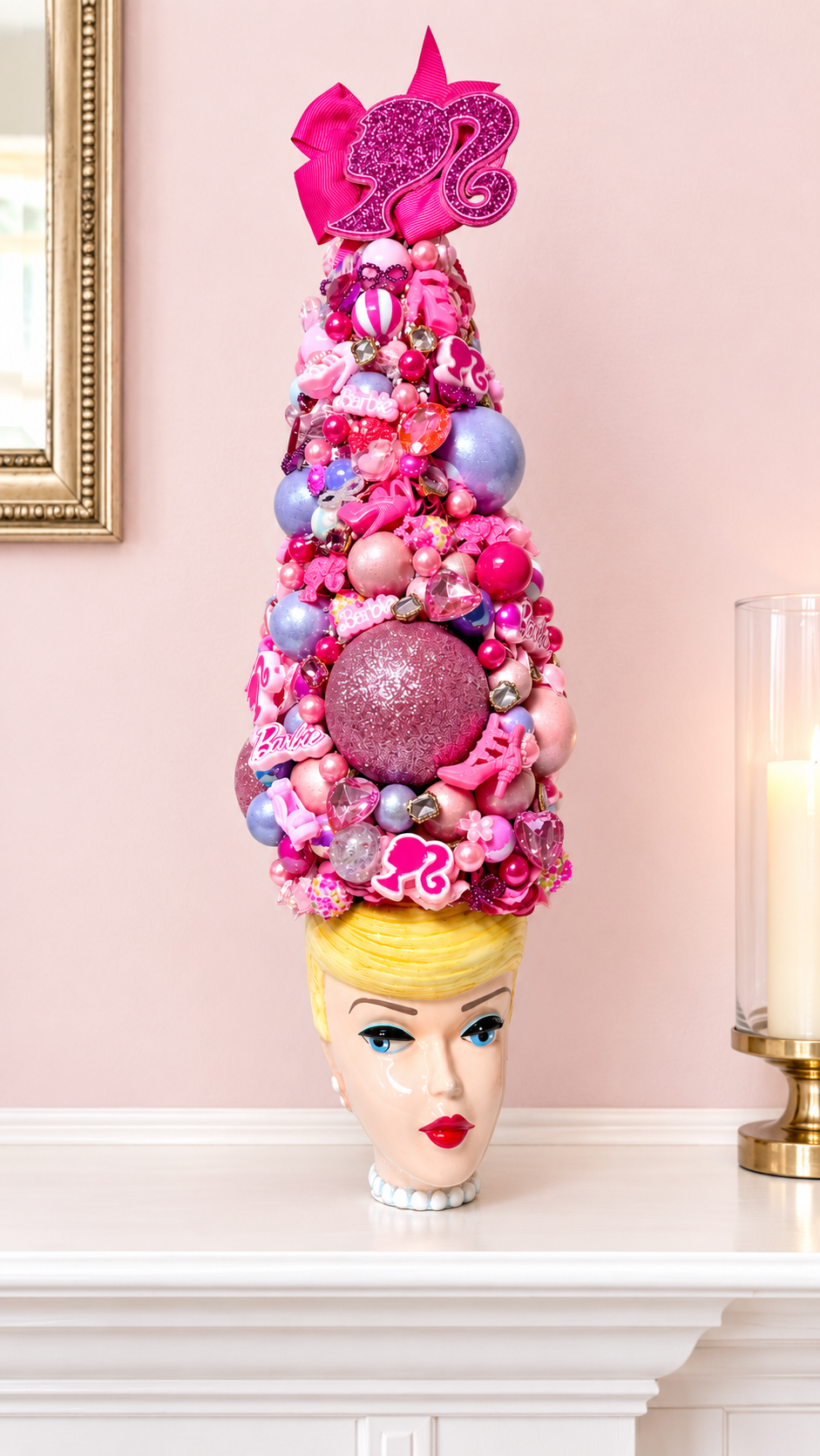 A decorative, surreal sculpture of a Barbie-themed Christmas tree on a mannequin head, set on a white mantelpiece against a soft pink wall.