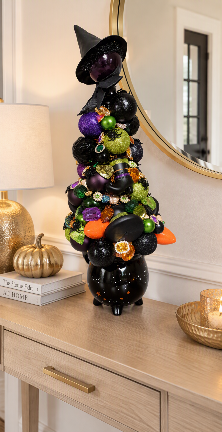 A decorative black tree made of stacked glossy black ornaments and topped with a witch hat, styled on a light wood console table in a home interior with a lamp, books, and mirror in the background.