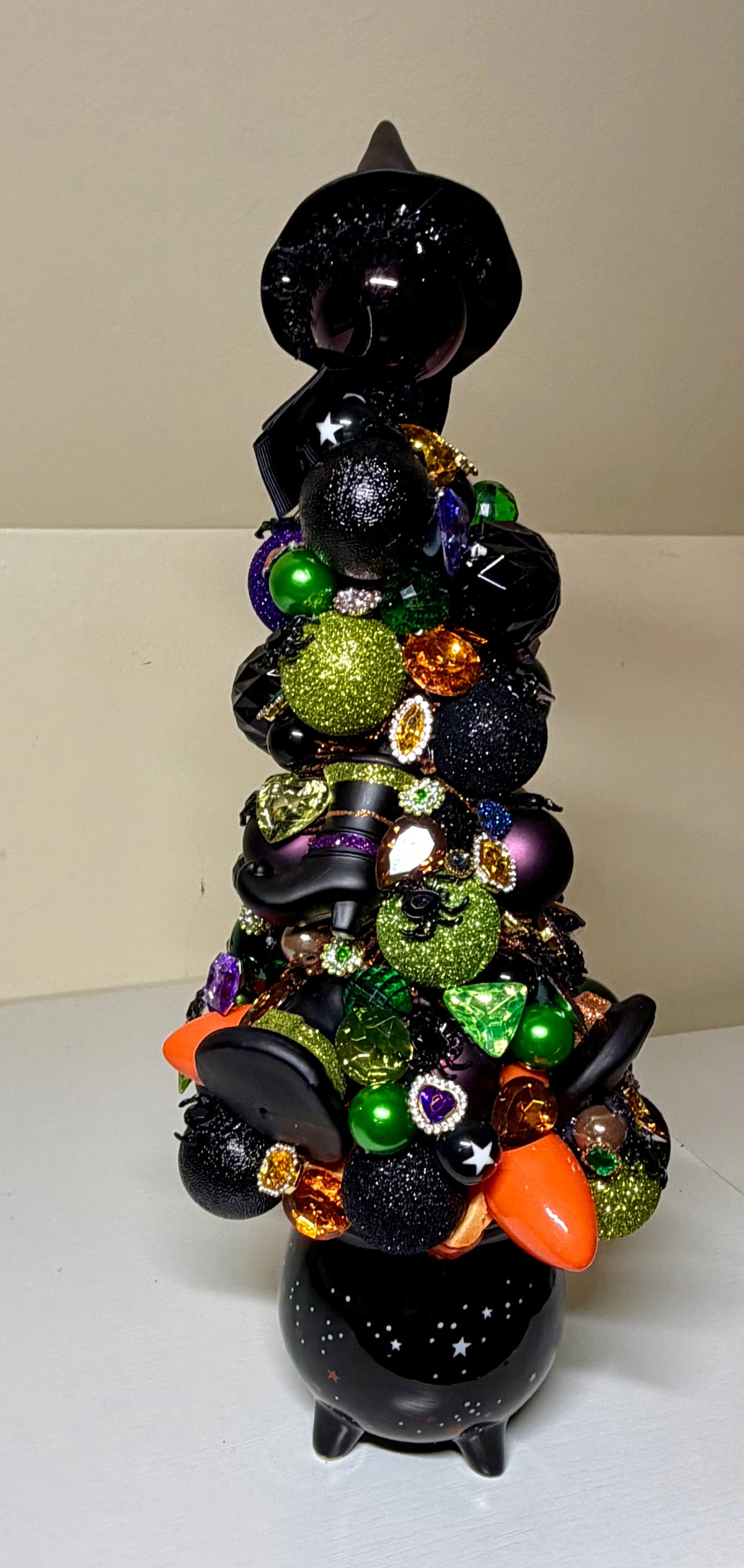 A black witch-themed decorative tree figurine adorned with purple, green, and orange ornaments, glittery accents, and small bat and pumpkin details, set against a plain beige background.