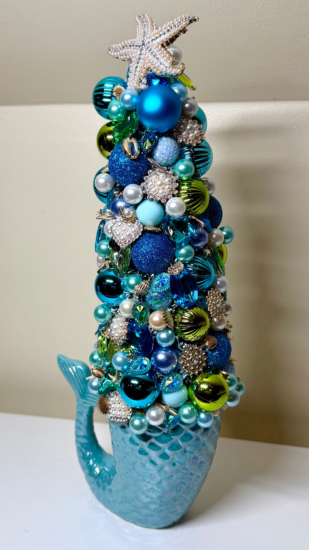 A decorative mermaid tail shaped Christmas tree ornament filled with blue, teal, green, silver, and white ornaments, topped with a glittery starfish, displayed on a white surface against a beige wall.