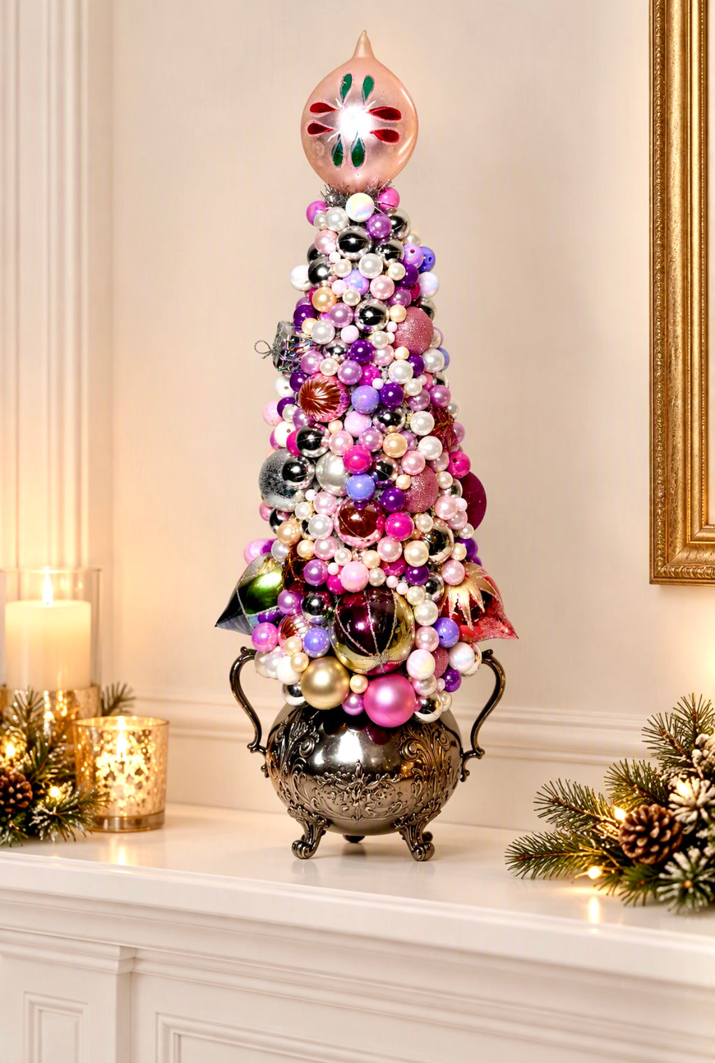 A decorative Christmas tree centerpiece with a metallic silver base, covered in colorful ornaments in pink, purple, white, red, green, and gold, topped with a pear-shaped ornament, displayed on a white mantelpiece with candles and holiday greenery.