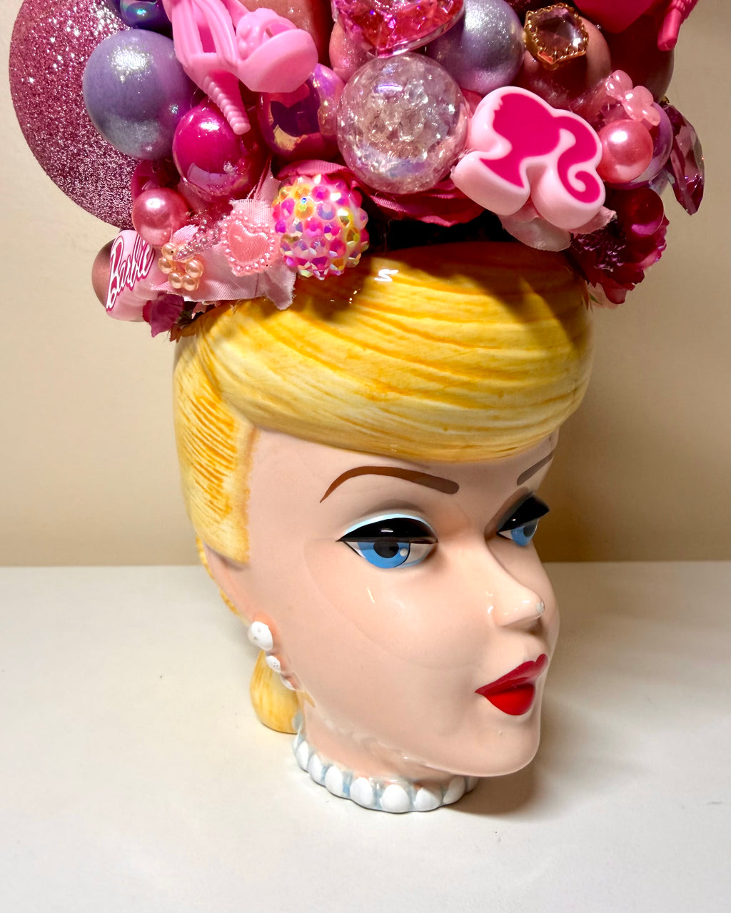 A ceramic doll head planter featuring a blonde character face with blue eyes and red lips, topped with a large pink and purple decorative headpiece, shown on a white surface against a beige background.
