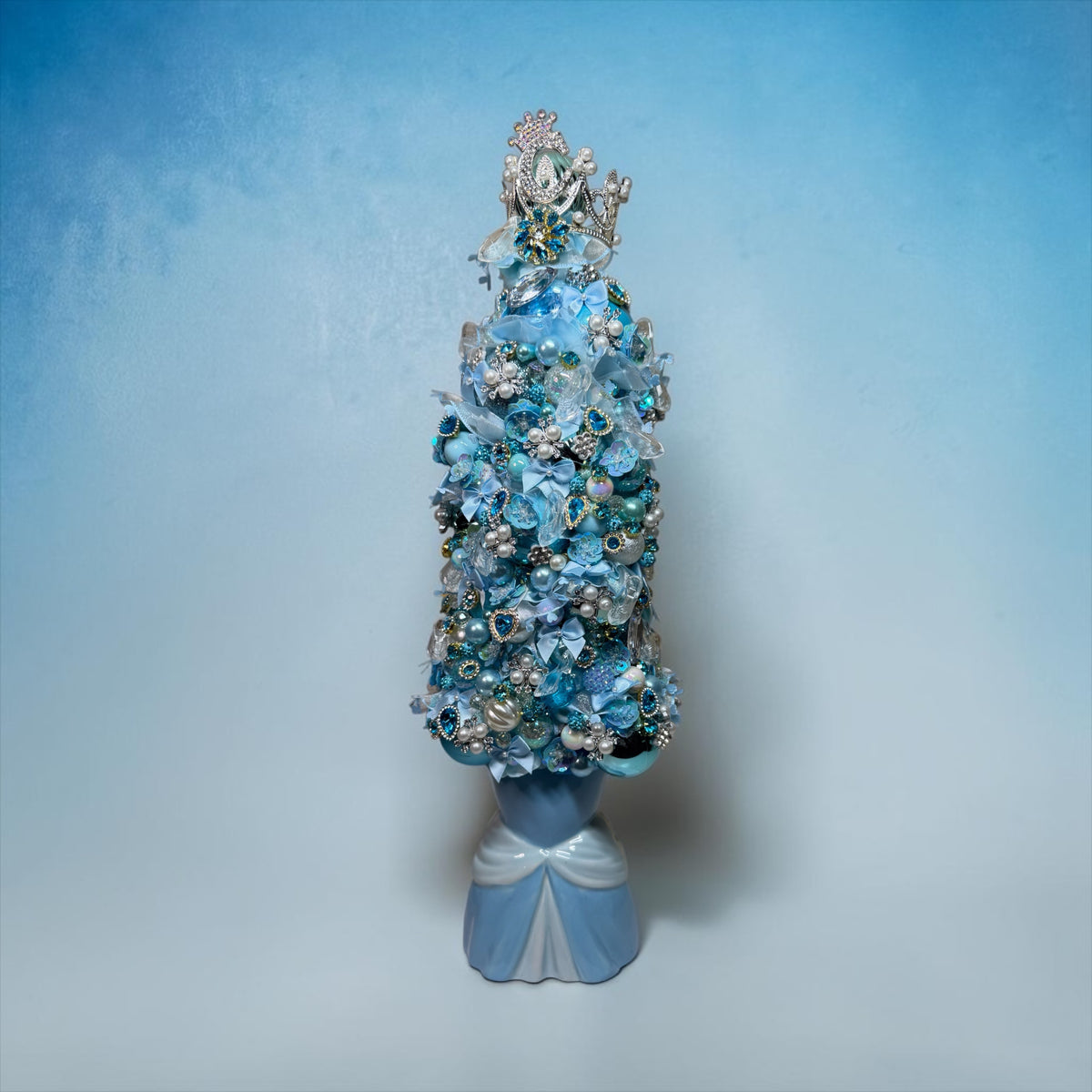 A highly embellished blue and silver floral crown displayed on a decorative blue stand against a soft blue gradient background.