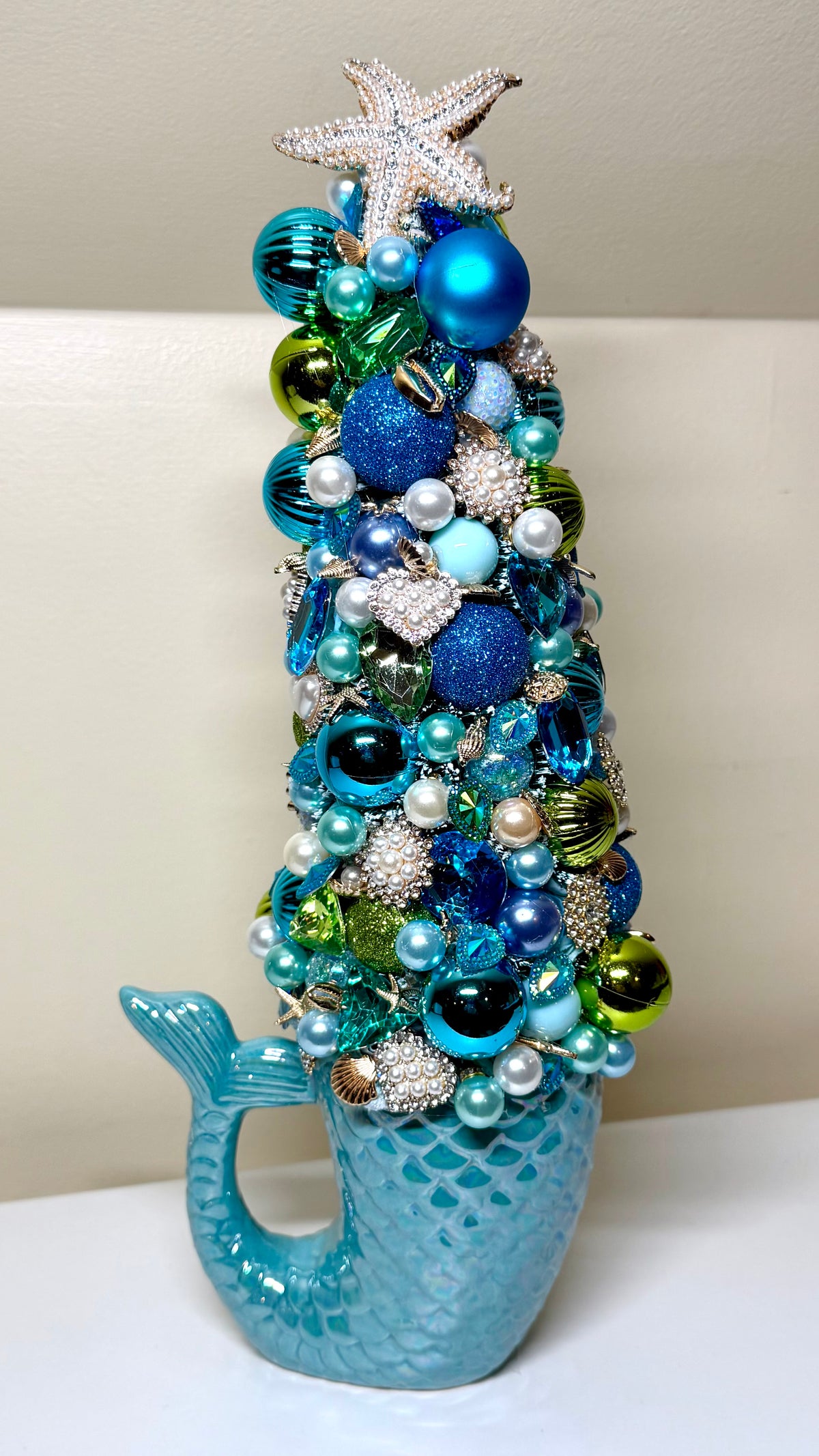 A decorative mermaid-themed Christmas tree ornament featuring a blue scaled whale tail base and a cone-shaped tree covered in blue, teal, green, and silver ornaments, topped with a silver starfish.