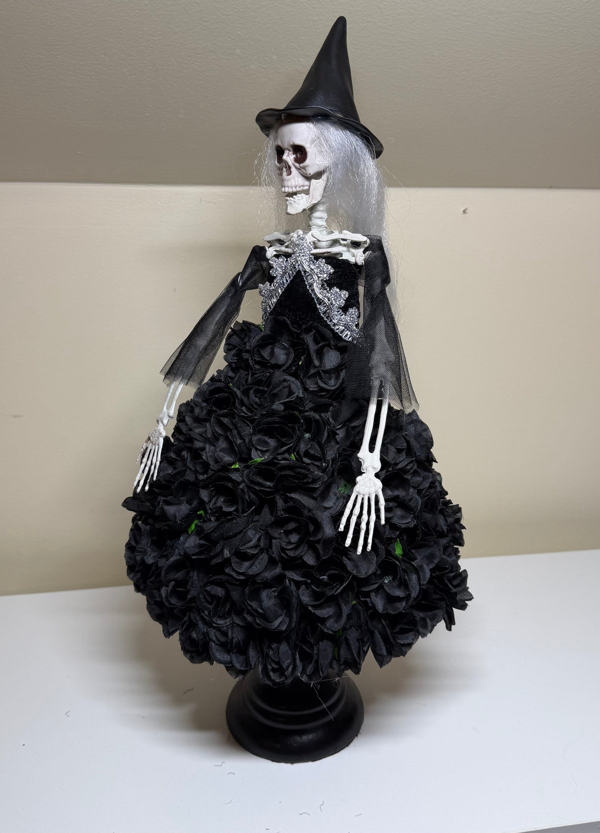 A Halloween skeleton witch decoration with a black pointed hat, white skull face, silver-gray hair, and a black rose-covered dress on a black stand, photographed indoors on a white shelf against a beige wall.