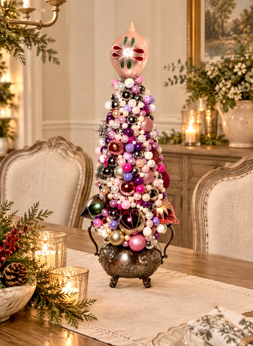A decorative tabletop Christmas tree in a silver footed pot, adorned with many colorful ornaments and beads, topped with a pink ornament, set on a dining table in a warmly lit room.