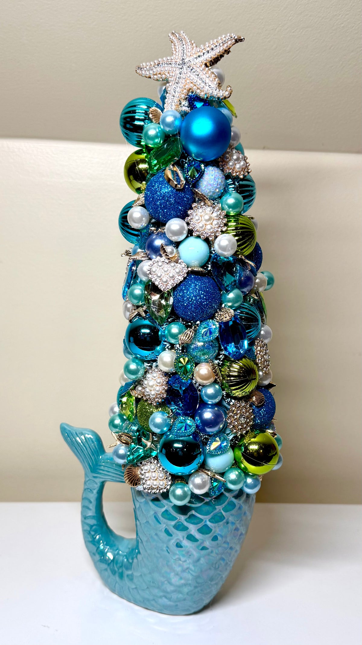 A decorative mermaid-tail-shaped Christmas tree ornament in shimmering turquoise blue, decorated with blue and teal baubles, pearl-like beads, glittery silver ornaments, and a silver starfish topper, shown against a neutral indoor background.