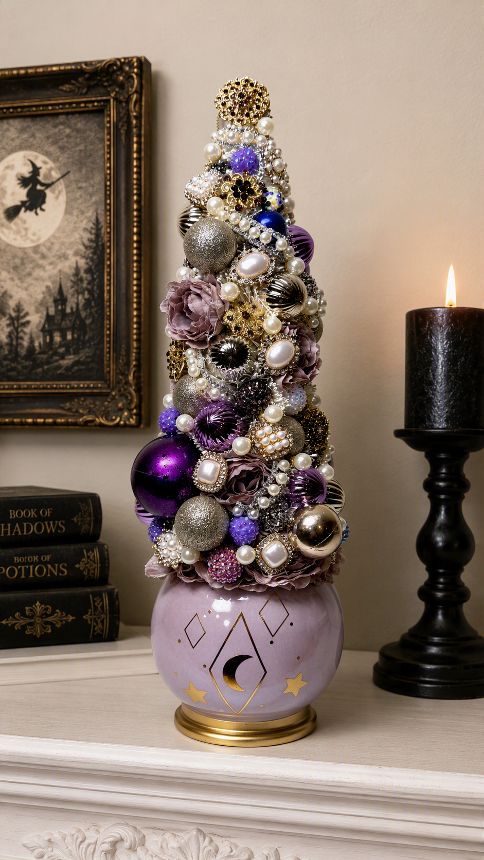 A decorative tabletop Christmas tree styled in a witch-themed setting, featuring purple and silver ornaments, faux pearls, and floral accents on a lavender ceramic base with gold details, set on a mantel next to a lit candle and framed artwork.