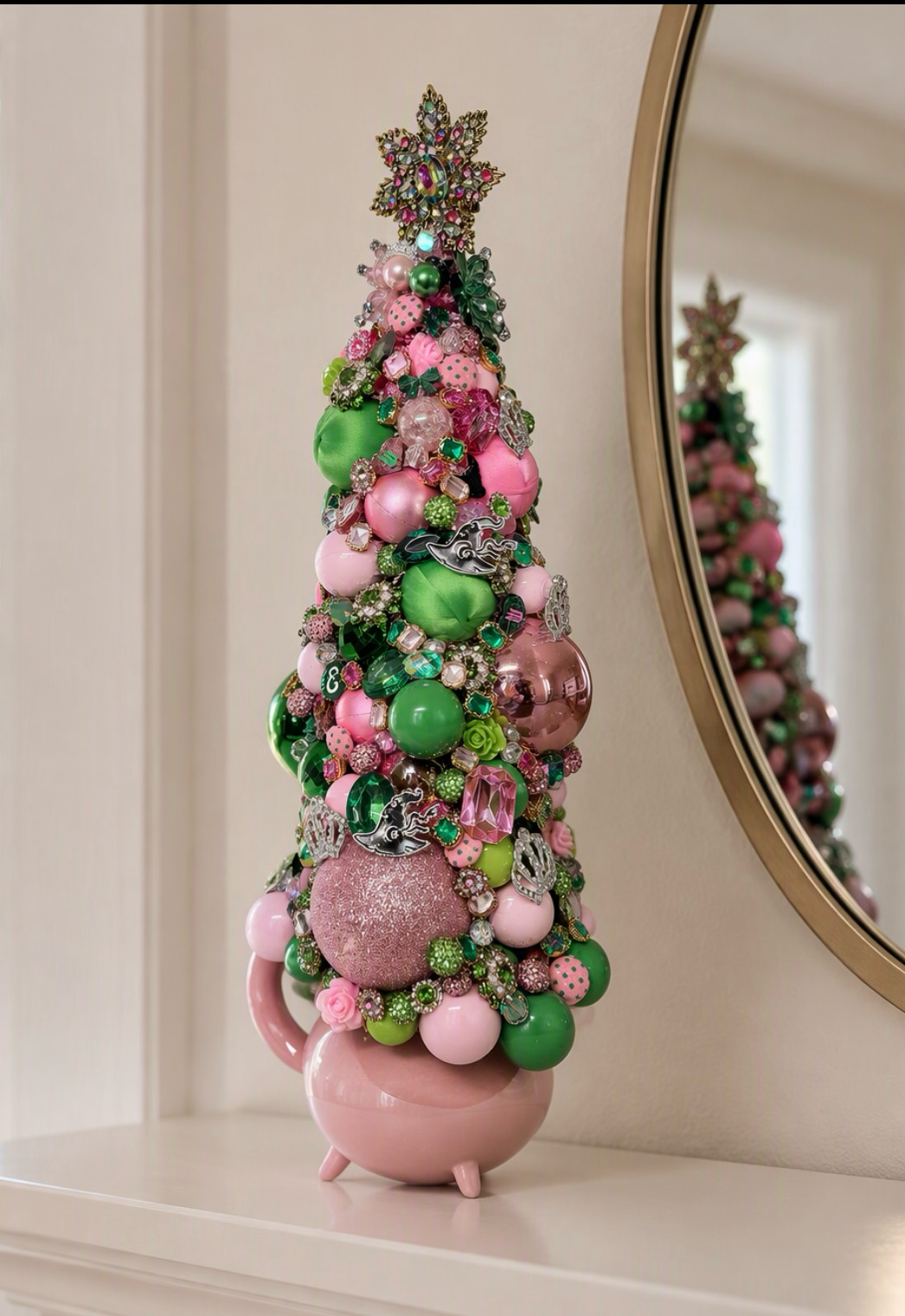 A decorative Christmas or Table Top tree made from pink and green ornaments, beads, and sequins, displayed on a white mantelpiece next to a round mirror.