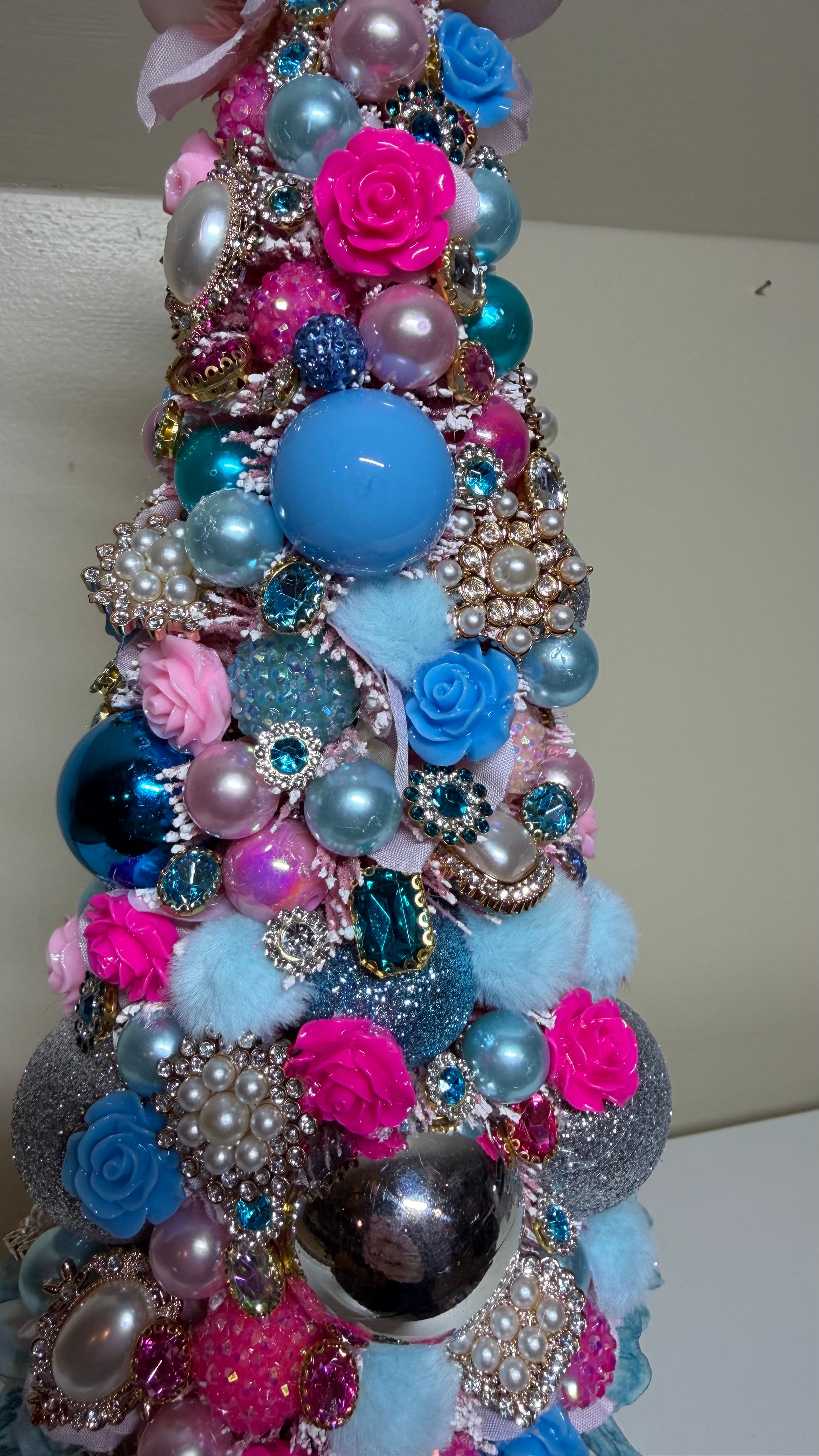 A decorative conical tree ornament covered in pink and blue faux flowers, glossy blue and light blue beads, pearl-like spheres, rhinestone brooch elements, glittery accents, and fluffy pom-pom details, photographed against a plain light background.