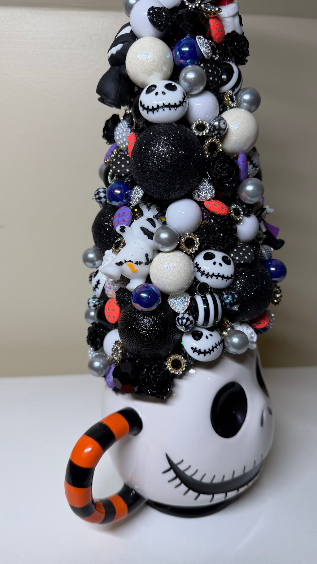 A decorative Halloween-themed mug shaped like a face, filled with a tall cluster of black, white, and silver ornaments, beads, and pom-poms, with an orange-and-black striped handle.