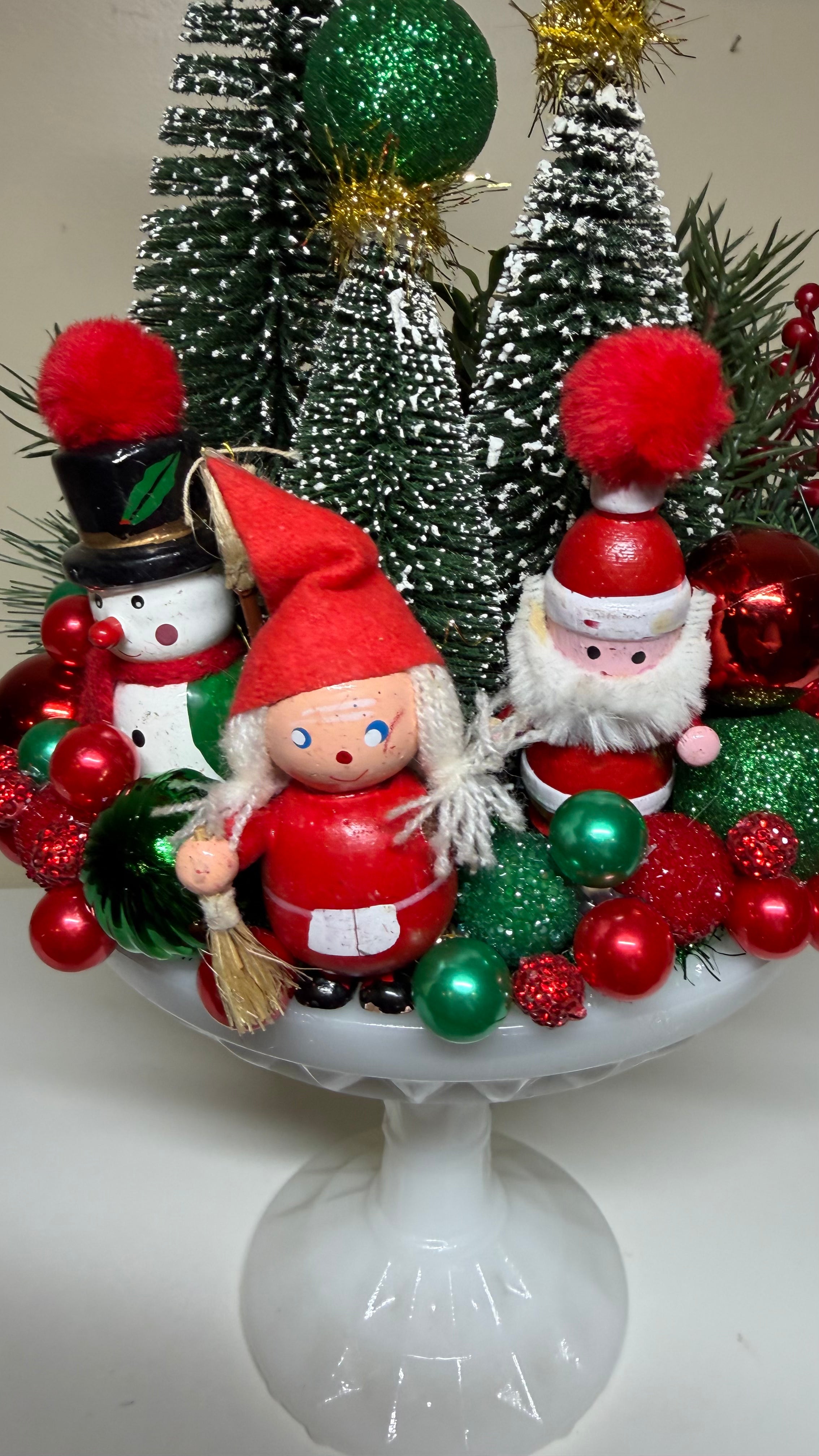 A festive holiday tabletop arrangement in a white pedestal bowl featuring a small wooden gnome in red clothing, a snowman ornament, a Santa figure, miniature green and white Christmas trees, red and green ornaments, and pine branches.