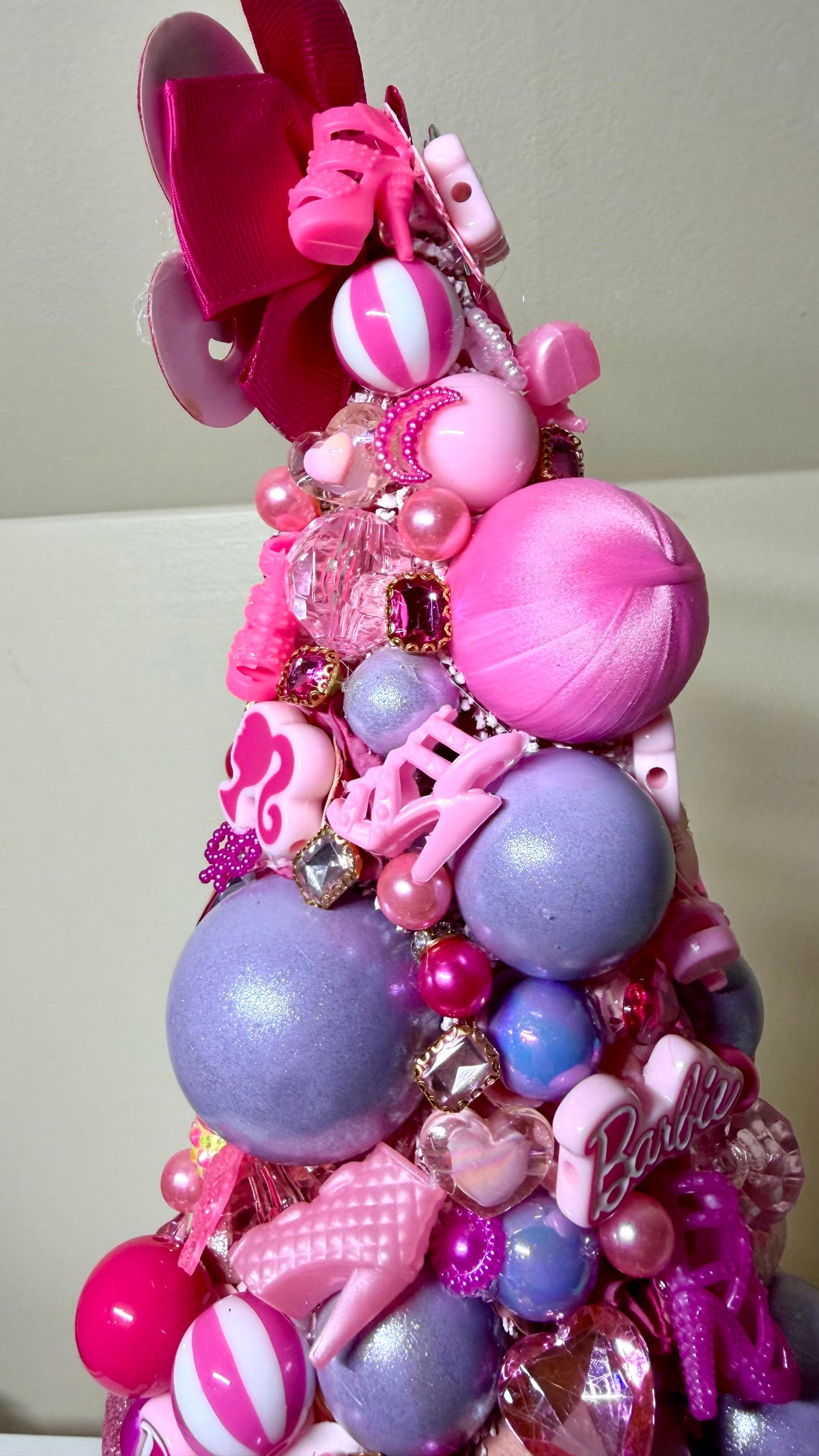 A pink and purple Barbie-themed decorative tree adorned with satin ornaments, pink bows, purple and pink balls, rhinestone accents, and a Barbie logo charm, photographed indoors against a light neutral background.