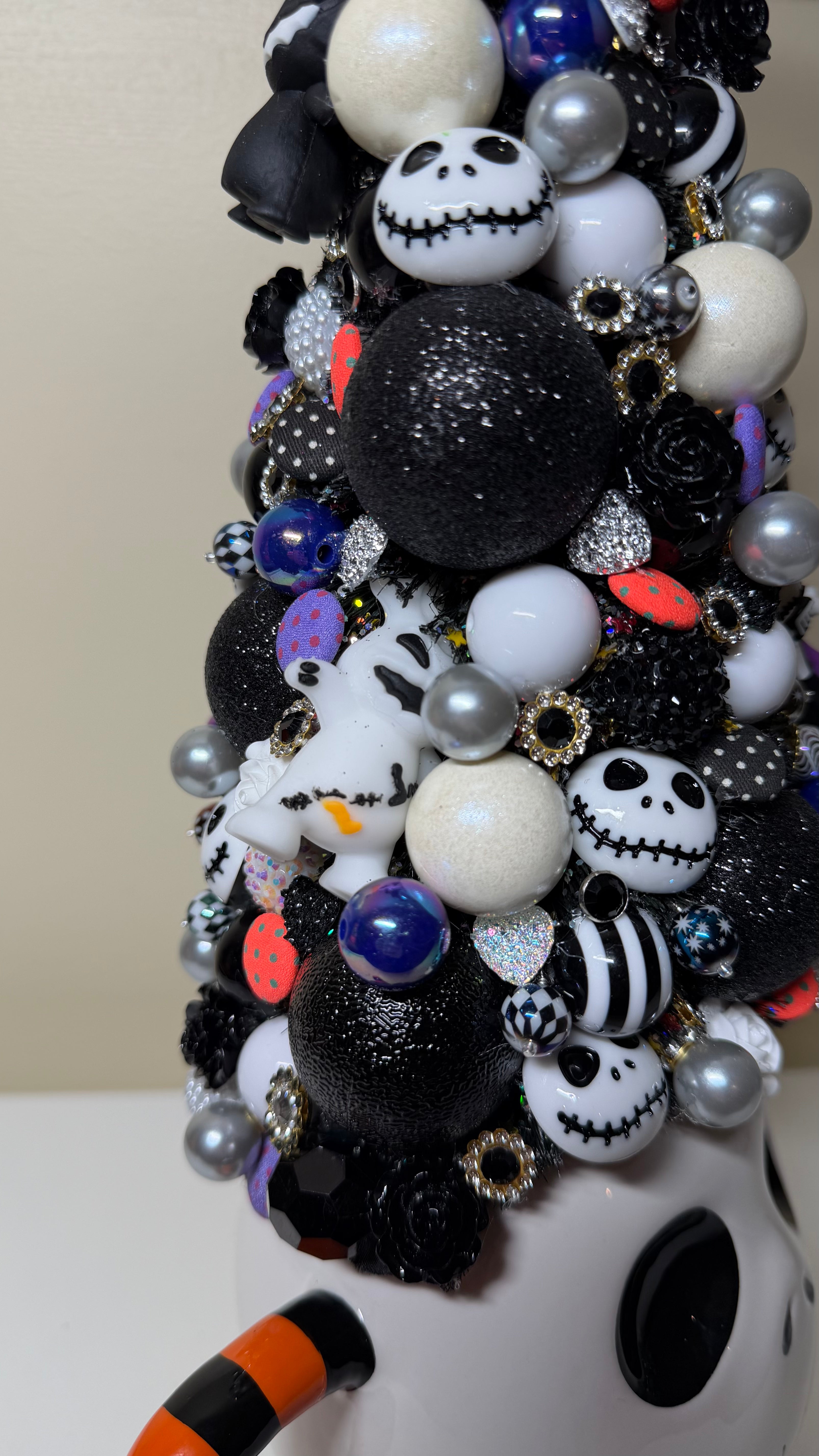 A close-up of a decorative Halloween ornament arrangement featuring black glitter balls, white and black Jack Skellington-themed ornaments, silver pearl-like beads, and small multicolor accents, mounted on a white surface.