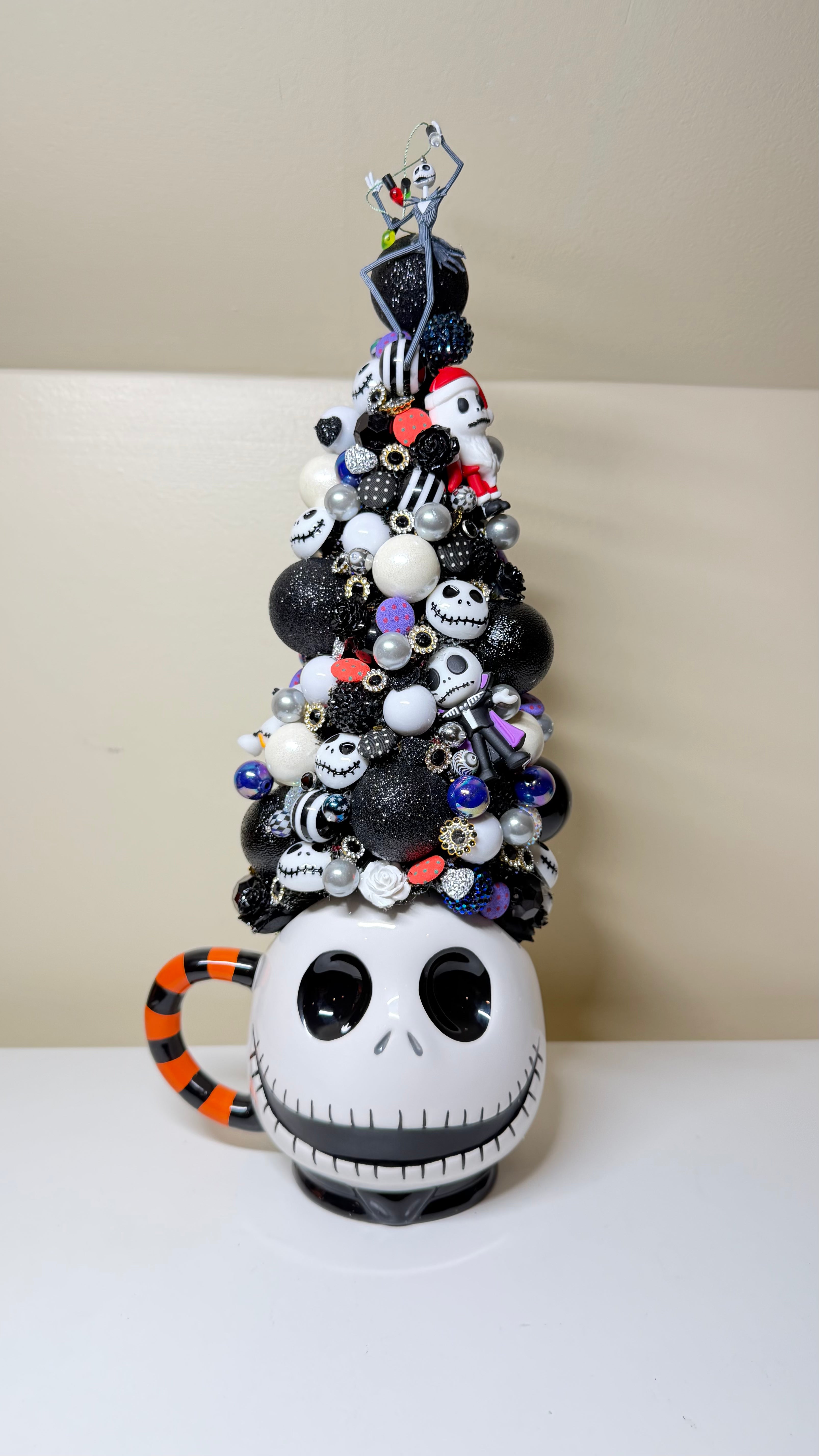A decorative Christmas tree crafted from a Jack Skellington mug, featuring black, white, and orange ornaments, with a small figure on top, set against a neutral beige background.
