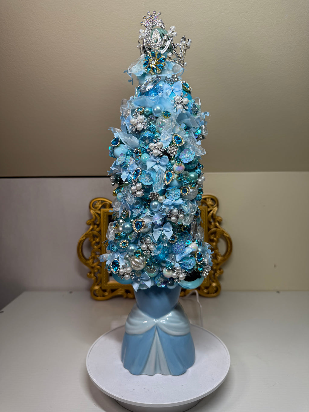 A decorative miniature Christmas tree in shades of light blue, white, and silver, styled with artificial flowers, pearls, and rhinestones, displayed on a white circular base against a beige wall with a gold frame behind it.