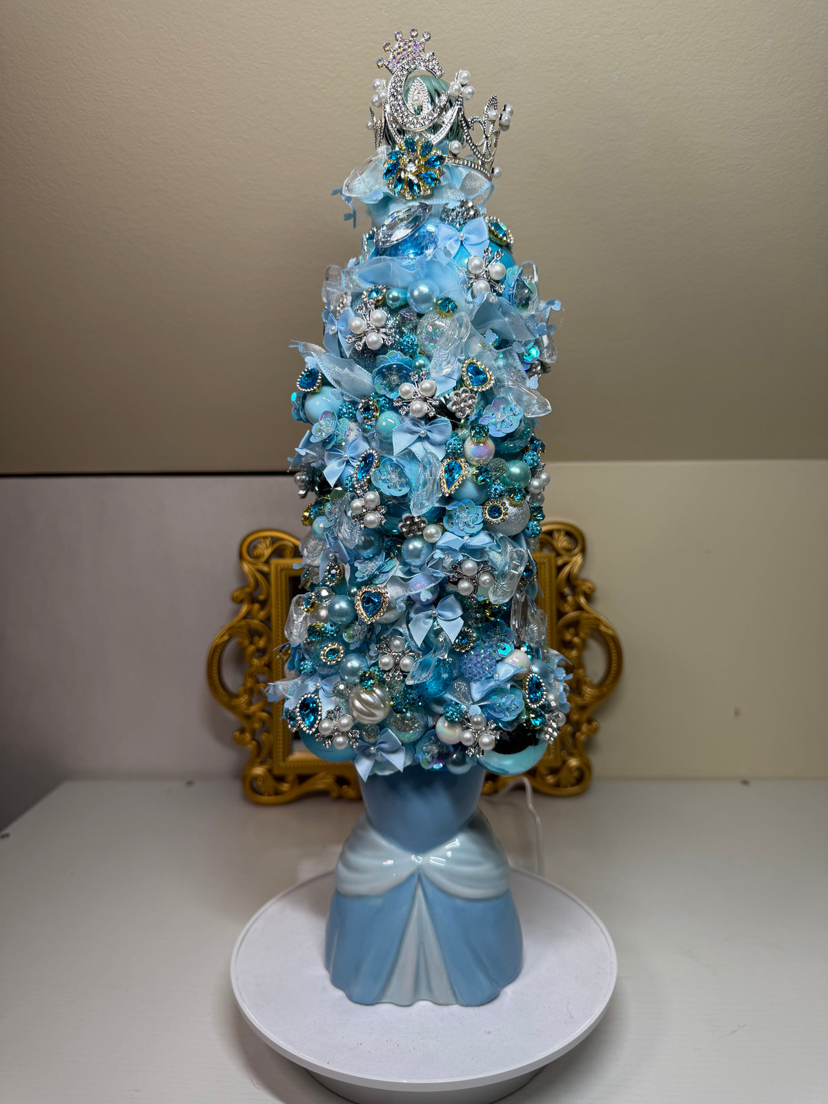 A decorative miniature Christmas tree in shades of light blue, white, and silver, styled with artificial flowers, pearls, and rhinestones, displayed on a white circular base against a beige wall with a gold frame behind it.