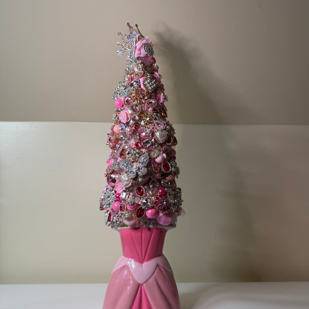 A pink and silver decorative Christmas tree covered in rhinestones, sequins, and pink ornaments, displayed on a pink dress-shaped stand against a beige wall.