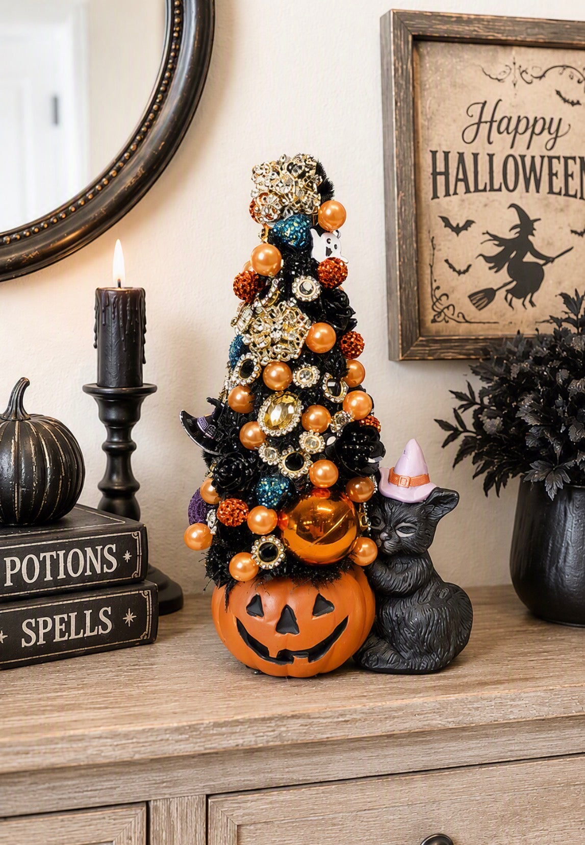 A decorative Halloween scene on a wooden dresser with a small black artificial tree ornament draped in orange and gold ornaments, a carved orange jack-o'-lantern base, a black cat figurine wearing a candy corn hat, candles, framed wall art, and a mirror.