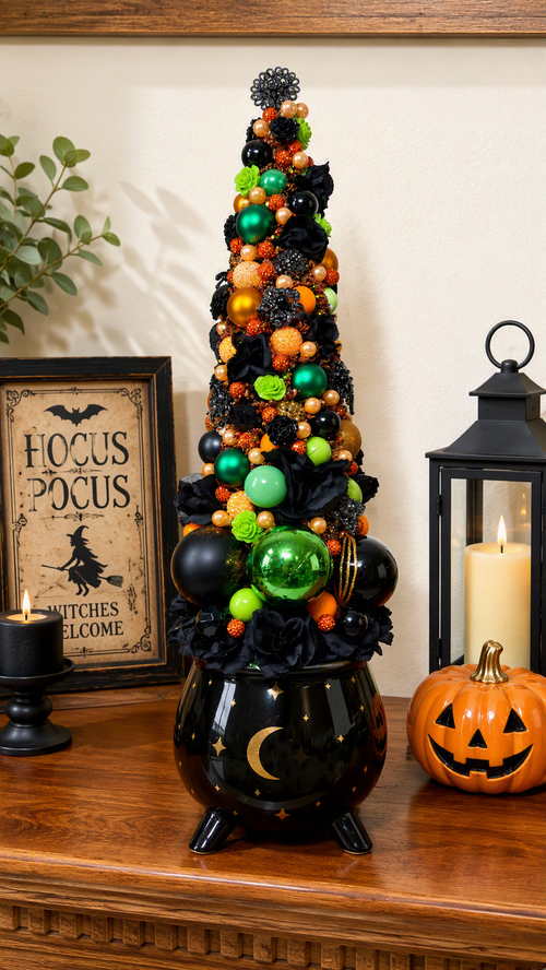 A Halloween-themed decorative tabletop centerpiece featuring a black cauldron-shaped base with a tall, narrow black tree adorned with green and orange ornaments, black fabric roses, and a black pom-pom topper. The centerpiece sits on a wooden mantel beside a framed 'Hocus Pocus' sign, a black candle holder, a black lantern with a lit candle, and a carved orange jack-o'-lantern.
