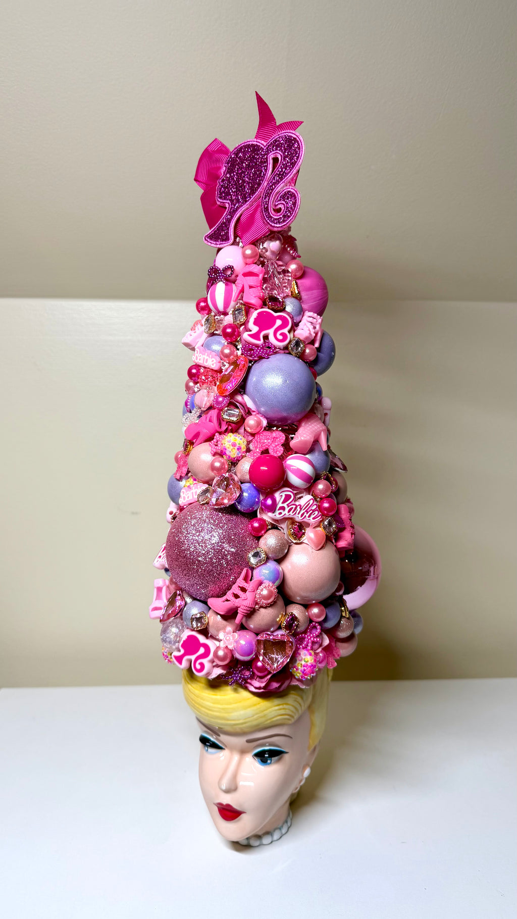 A Barbie-themed decorative hat or headpiece made of pink and purple ornaments, beads, and glitter, displayed on a blonde mannequin head against a plain beige background.