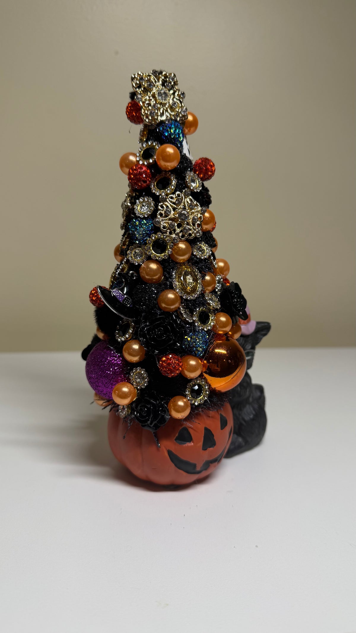 A Halloween-themed decorative tree figurine made from a black Christmas tree adorned with orange, gold, and purple beads, ornaments, and glitter, sitting on top of a carved orange pumpkin. A small black cat figurine is tucked behind the tree.