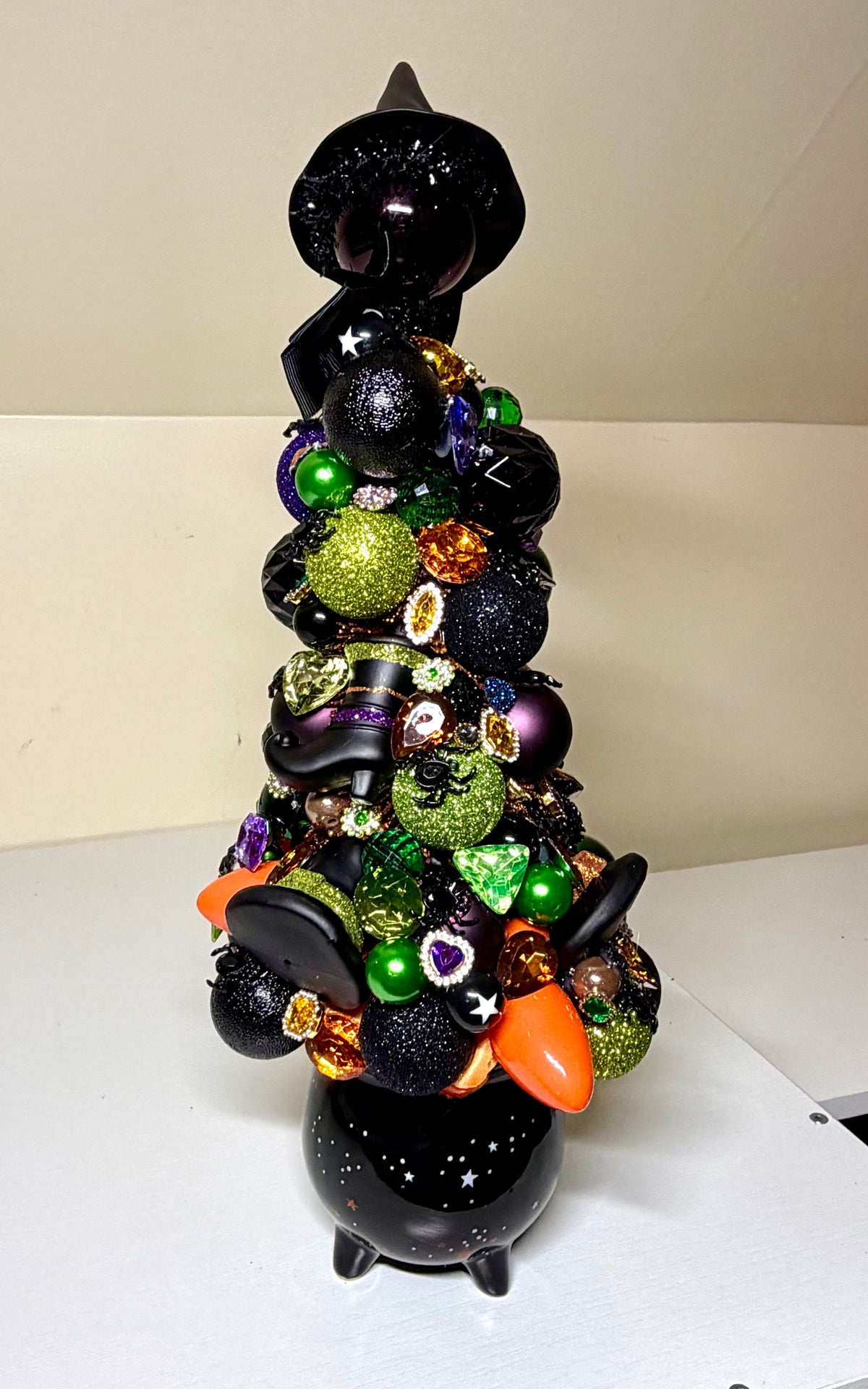 A black witch-themed Christmas tree decoration made from stacked black ornaments, glittery green ornaments, purple and orange accents, and small star details, displayed on a white surface against a plain beige wall.