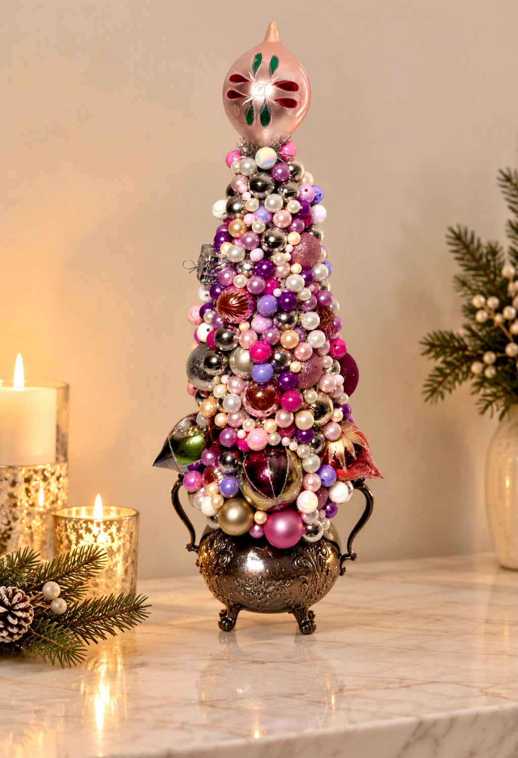 A decorative miniature Christmas tree ornament with a pink finial and a metallic bowl base, covered in colorful glass ornaments and beads, displayed on a marble surface with candles and greenery in a warm indoor setting.