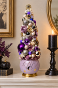 A decorative miniature Christmas tree with purple, silver, and gold ornaments, flowers, and faux pearls, displayed on a mantelpiece next to a lit black candle and a small black pumpkin.