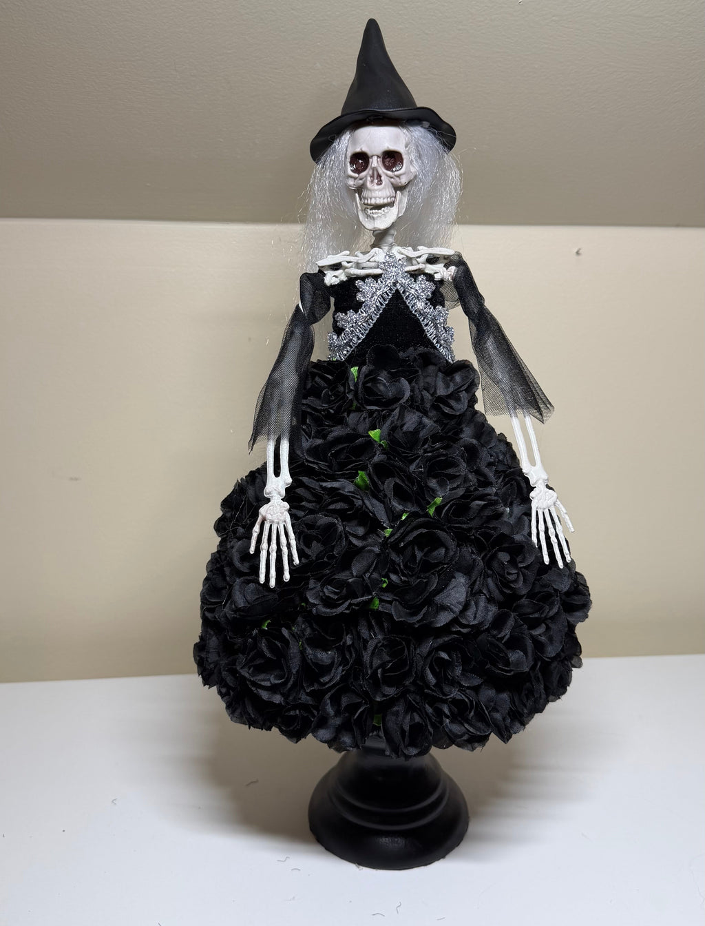 A Halloween skeleton witch decoration wearing a black pointed hat, black dress with gray lace details, and a black ruffled skirt on a black stand, displayed on a white shelf against a beige wall.