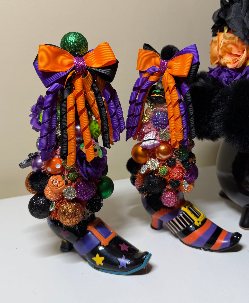 Two Halloween-themed witch hat boot decorations decorated with orange and purple bows, ribbons, pom-poms, and small orange jack-o'-lanterns, displayed on a white surface against a beige wall.