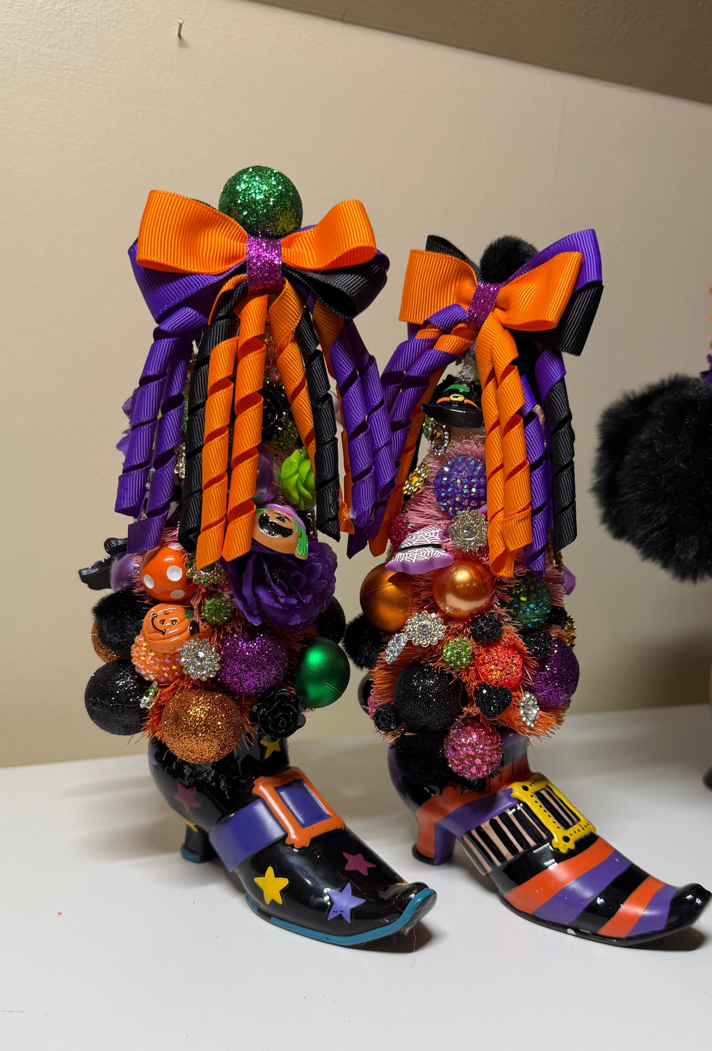 A pair of Halloween witch boot decorations on a white tabletop against a beige wall. The boots are black with orange and purple stripes, large orange and black bows, colorful glitter ornaments, and small jack-o'-lantern details.