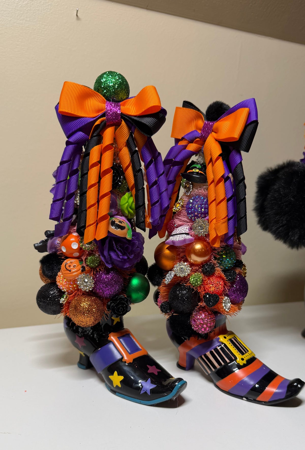 A pair of Halloween witch boot decorations on a white tabletop against a beige wall. The boots are black with orange and purple stripes, large orange and black bows, colorful glitter ornaments, and small jack-o'-lantern details.