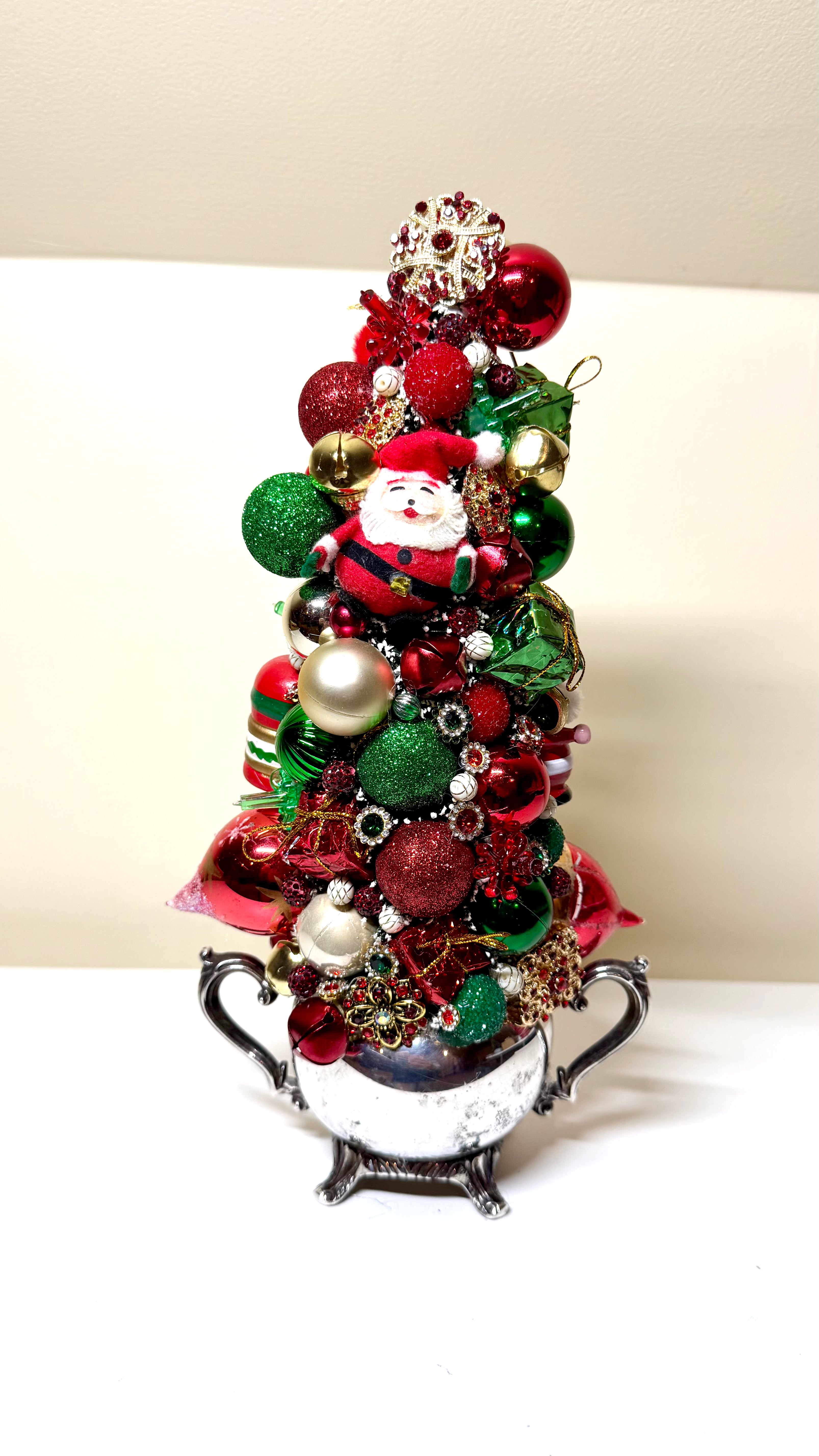 A decorative miniature Christmas tree arrangement in a white ceramic pot with black handles, decorated with red and green ornaments, shiny baubles, a Santa Claus figure, and a gold snowflake topper, set against a plain light background.