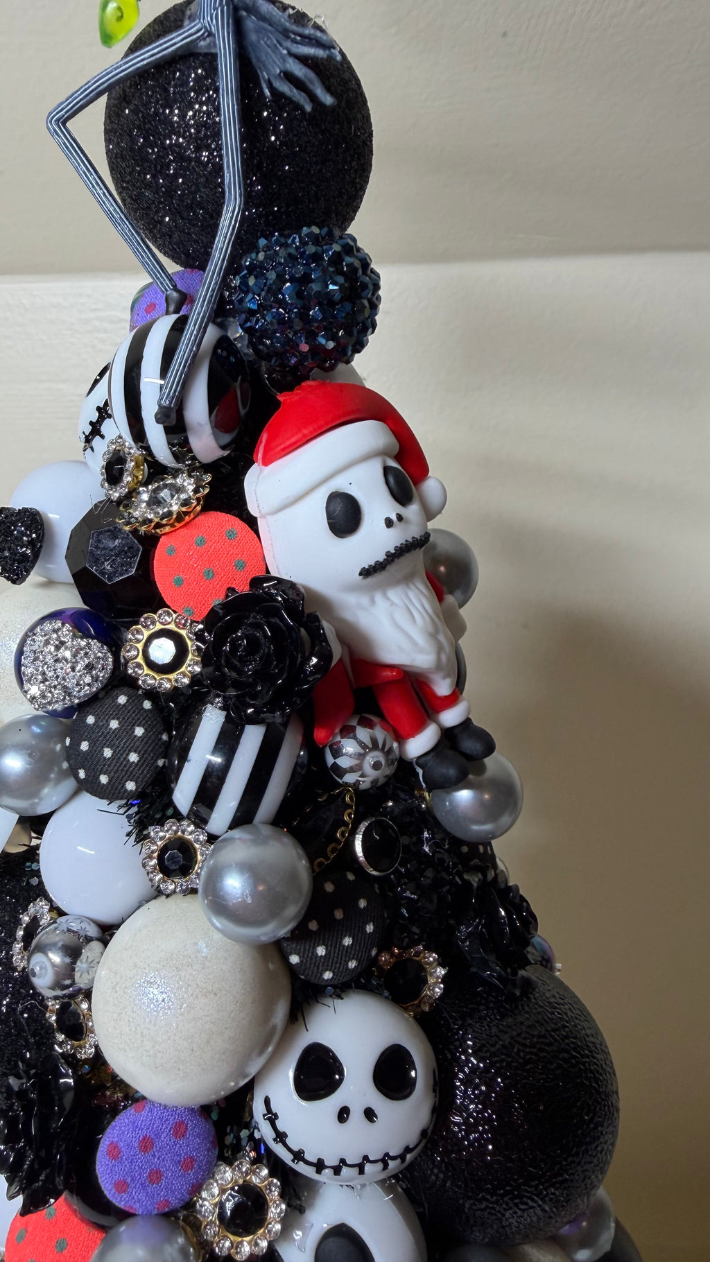 A decorative black Christmas tree adorned with various ornaments, including a white skull-shaped ornament, a red Santa hat figure, black and white striped balls, glittery pom-poms, and silver beads, set against a plain beige wall.