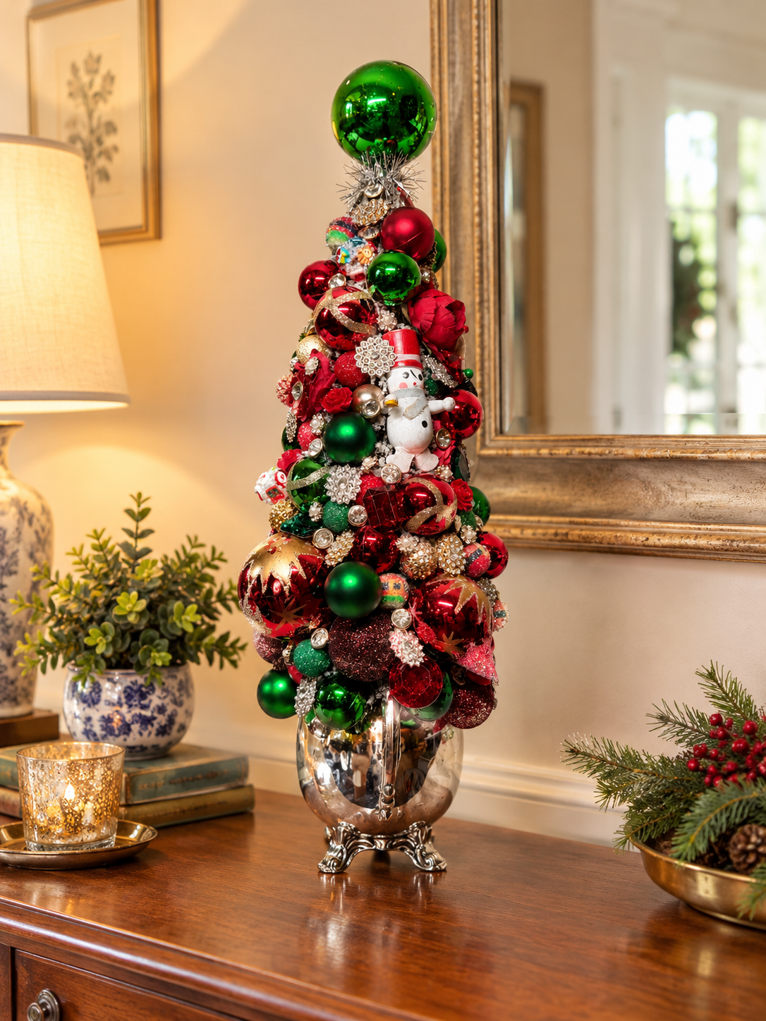 A decorative miniature Christmas tree in a silver footed pot, adorned with red and green ornaments, ribbons, snowflake ornaments, and a snowman figure, displayed on a wooden console table in a warmly lit room.